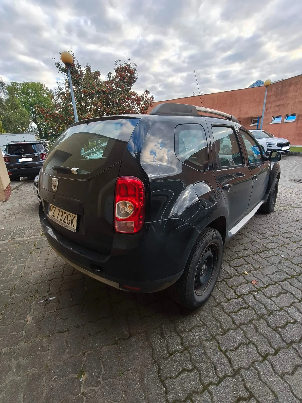 Czarny samochód marki Dacia Duster zaparkowany tyłem na kostce brukowej. Widoczna jest tablica rejestracyjna o numerze PZ 732GK. W tle znajduje się inny ciemny samochód, brązowy budynek oraz drzewa pod zachmurzonym niebem.