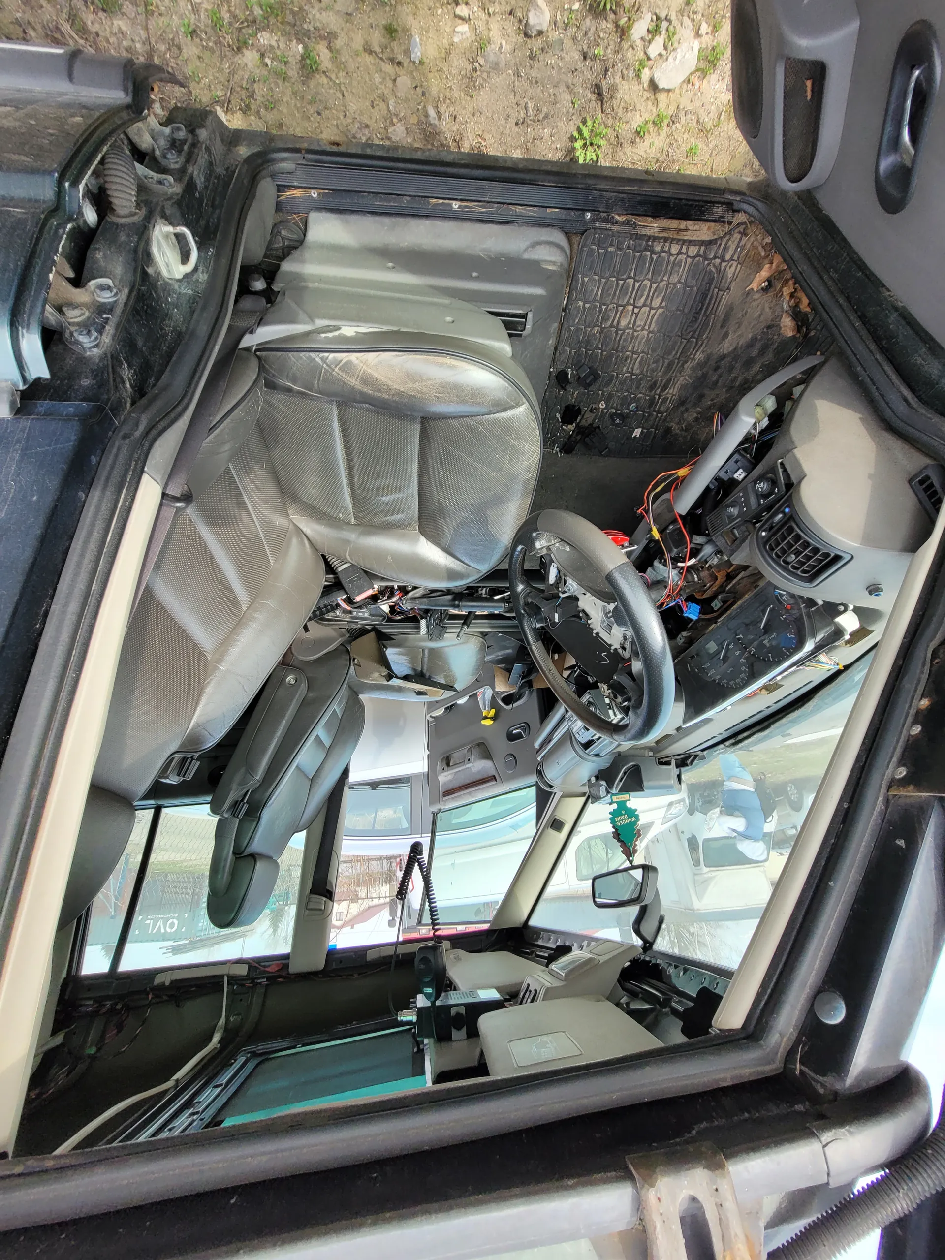 Wnętrze pojazdu z siedzeniami, kierownicą, deską rozdzielczą z odsłoniętymi przewodami i zielonym zapachem w kształcie choinki na lusterku wstecznym. Przez przednią szybę widoczne są inne pojazdy oraz budynek z napisem "OVL". Podłoga pokryta czarną wykładziną, po prawej stronie obrazu fragment ziemi z kamieniami i roślinami.