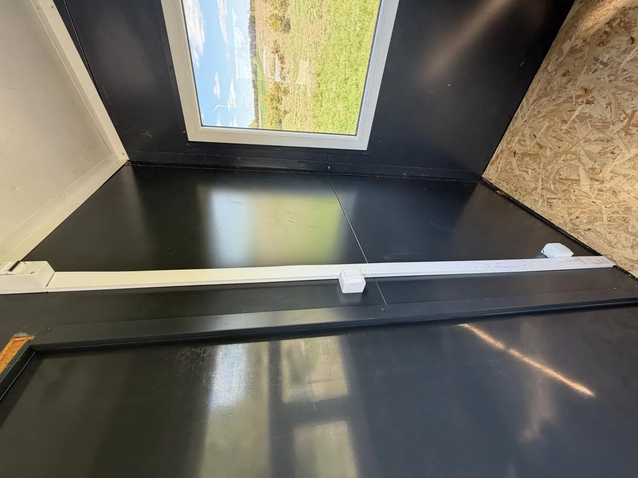 Wnętrze struktury z czarną podłogą, białą ścianą po lewej stronie i ścianą z płyty OSB po prawej. W tle widoczne okno lub skylight pokazujące zielone pole i niebo z chmurami. Na czarnej powierzchni umieszczony jest biały poziomy element z małym białym pudełkiem lub czujnikiem.