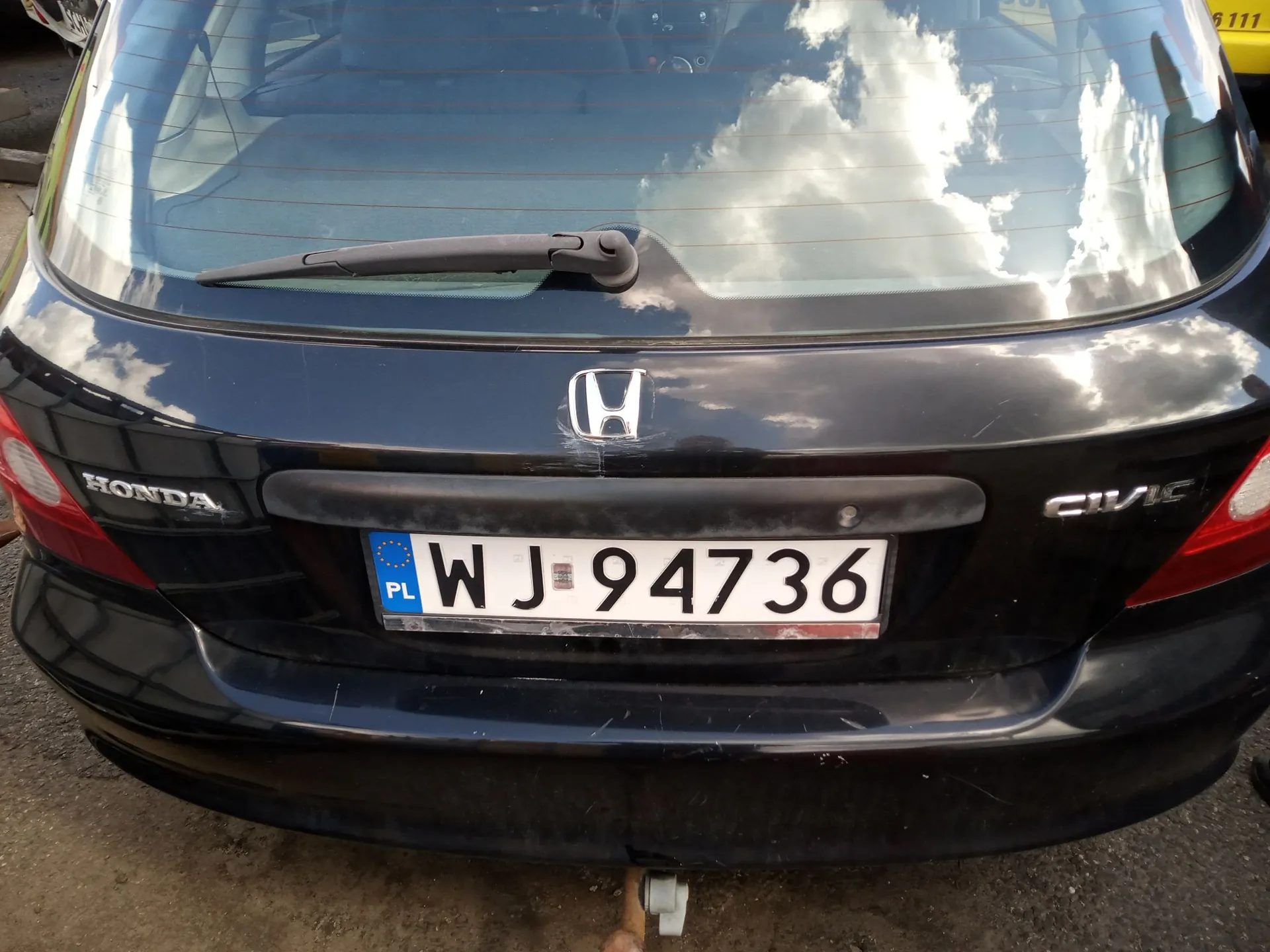 Tylny widok czarnego samochodu marki Honda Civic. Na klapie bagażnika znajduje się logo Hondy oraz napisy 'HONDA' po lewej i 'CIVIC' po prawej stronie. Samochód posiada polską tablicę rejestracyjną o numerze WJ 94736. W tylnej szybie odbijają się chmury i niebo.