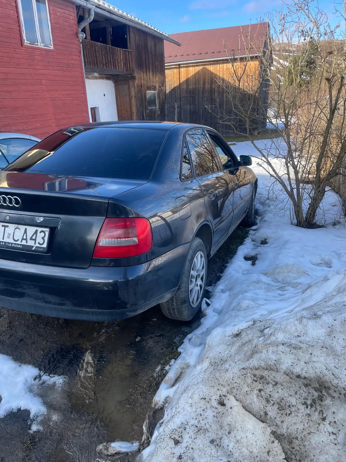 Czarny samochód Audi z tablicą rejestracyjną T CA43 zaparkowany na błotnistej drodze obok śnieżnych zalegów. W tle czerwony i drewniany budynek, nagie drzewa oraz niebieskie niebo. Śnieg częściowo roztopiony, widoczne ślady opon.