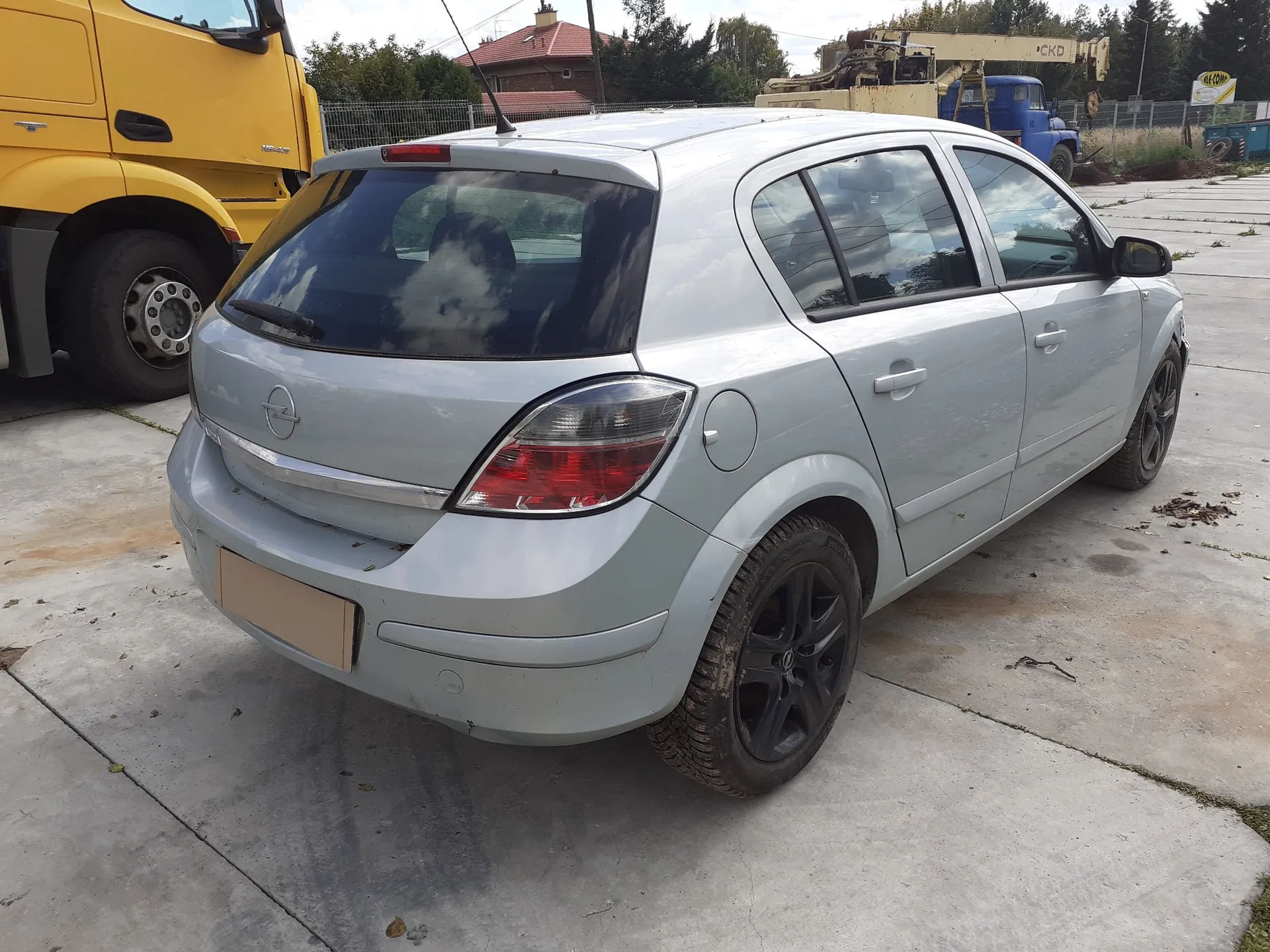 Srebrny samochód osobowy marki Opel Astra w nadwoziu hatchback, zaparkowany tyłem do widza na betonowym placu. Po lewej stronie widoczny jest fragment żółtej ciężarówki. W tle znajdują się drzewa, budynek z czerwonym dachem oraz niebieska maszyna budowlana.