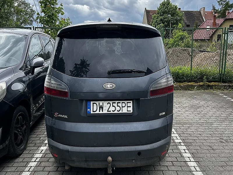 Zdjęcie przedstawia tył ciemnoszarego samochodu marki Ford S-MAX zaparkowanego na brukowanym parkingu. Widoczna jest tablica rejestracyjna z numerem DW 255PE oraz oznaczeniem PL. Po lewej stronie częściowo widoczny jest czarny samochód. W tle zielone ogrodzenie, krzewy, domy z czerwonymi dachami i chmurne niebo.