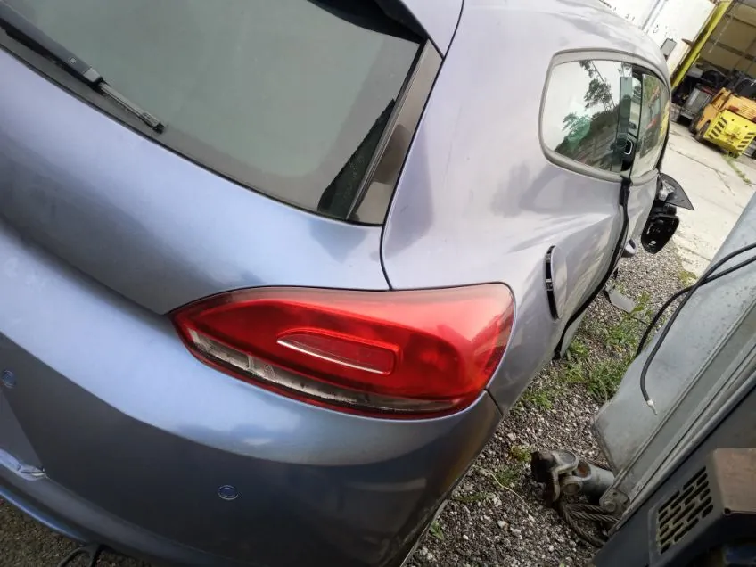 Zbliżenie na tylną część srebrnego samochodu. Widoczny czerwony tylny reflektor, boczne lusterko w prawej części kadru oraz fragment tylnej szyby z wycieraczką. Samochód stoi na podłożu z drobnej kostki i żwiru. W tle po prawej stronie widoczne są żółte elementy maszyn lub urządzeń oraz metalowa konstrukcja z przewodami. Na ziemi leżą również narzędzia lub części mechaniczne.