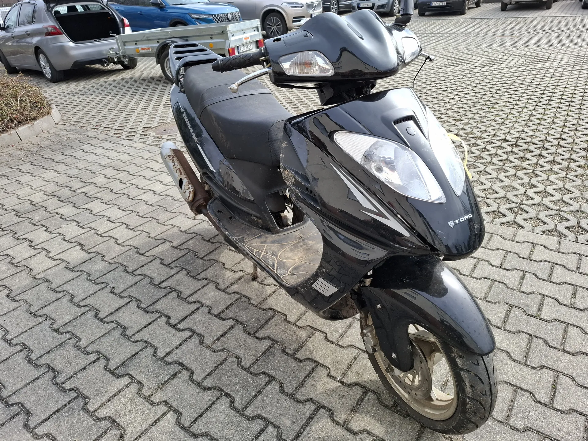 Czarny skuter marki TORO zaparkowany na chodniku z betonowych płyt. Skuter ma widoczne ślady zużycia, rdzę na wydechu i zarysowany napis TORO na przodzie. W tle stoją samochody, w tym srebrny z otwartym bagażnikiem i niebieski z przyczepą. Chodnik składa się z szarych, geometrycznych płyt.