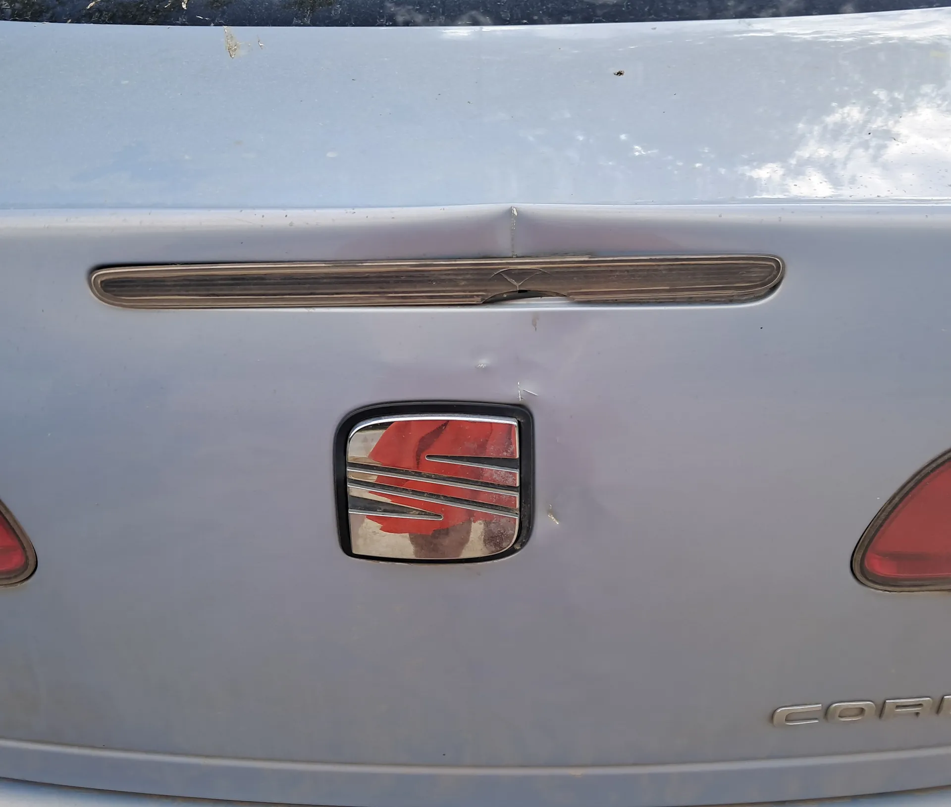 Zbliżenie na tylną część srebrnego samochodu. W centrum znajduje się emblemat marki SEAT z czerwonym tłem i srebrnymi pionowymi liniami. Nad emblematem widoczna jest pozioma, chromowana listwa z uszkodzeniem w środkowej części. Po obu stronach emblematu częściowo widoczne są czerwone światła tylnego. W prawym dolnym rogu widoczny jest fragment napisu 'COR' wykonanego srebrnymi literami. Na powierzchni nadwozia widoczne są drobne zadrapania i zabrudzenia.