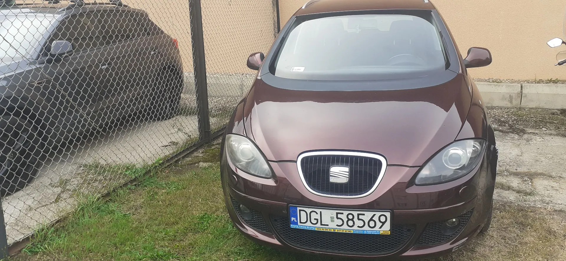 Widok z przodu brązowego samochodu marki SEAT zaparkowanego obok metalowego ogrodzenia z siatki. Samochód ma polską tablicę rejestracyjną o numerze DGL 58569. Po lewej stronie, za ogrodzeniem, widoczny jest fragment innego, ciemnego samochodu.