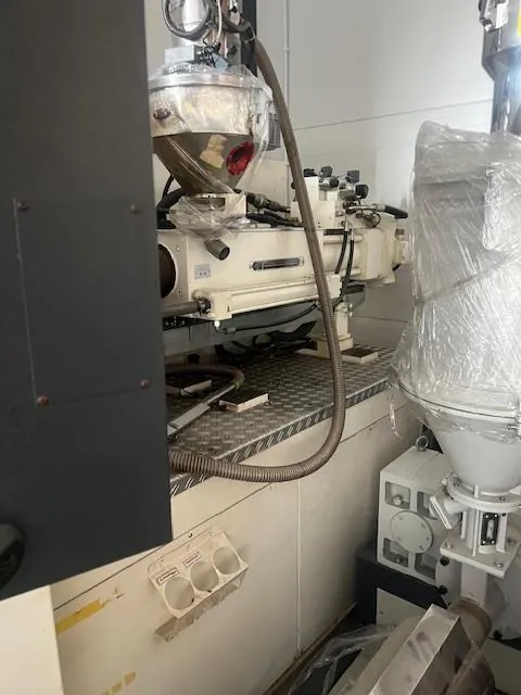 Przemysłowe urządzenia mechaniczne w pomieszczeniu. W centrum biała maszyna z przezroczystym zbiornikiem na górze, częściowo owiniętym folią. Do maszyny podłączone są grube węże. Poniżej znajduje się metalowa powierzchnia z wzorem rombów. Po prawej stronie drugi aparat z białym zbiornikiem, również owiniętym folią. Na ścianie pod maszyną biały panel z trzema okrągłymi otworami. Tło stanowi biała ściana. Brak czytelnego tekstu.