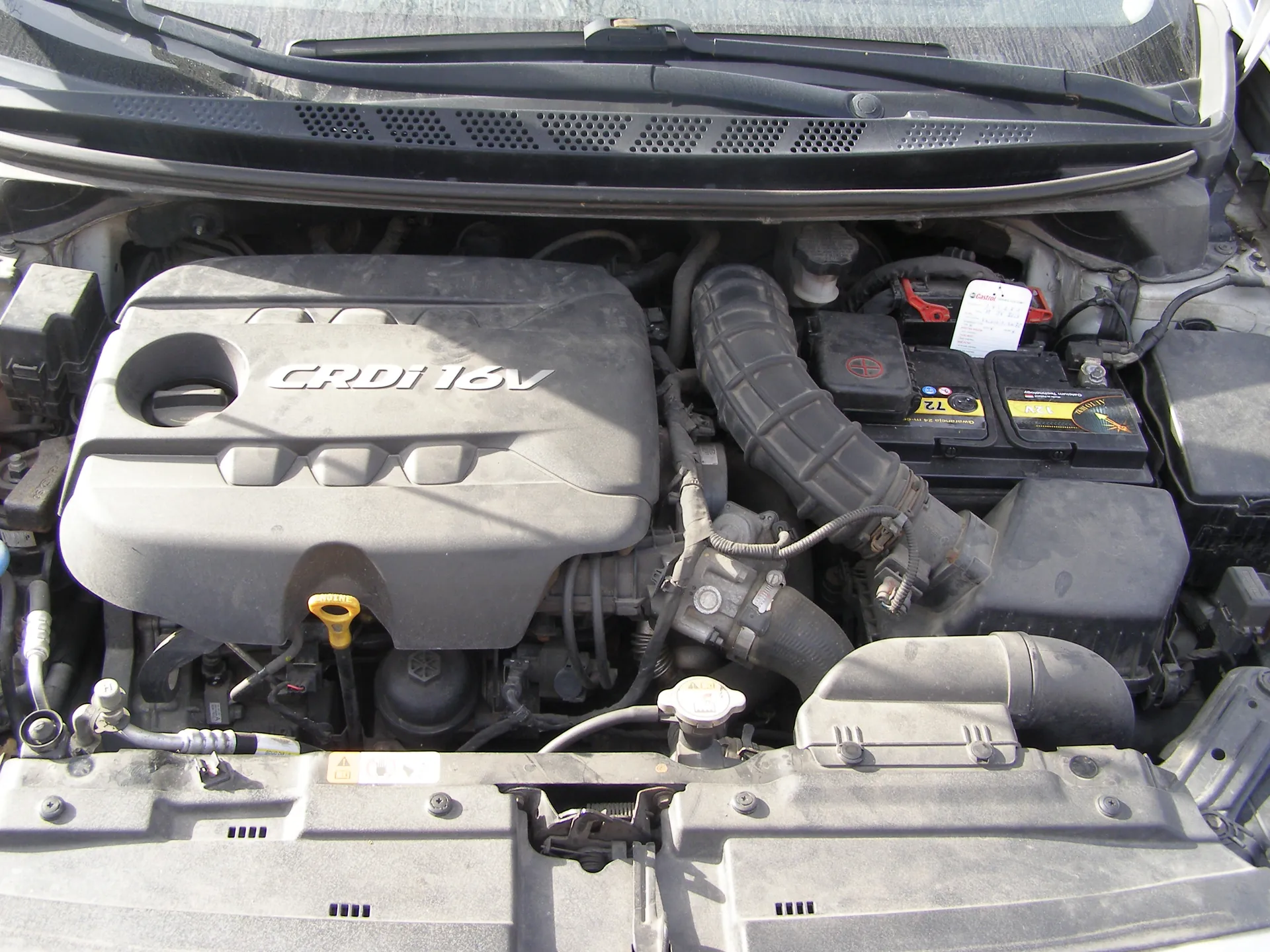 Widok otwartej tylnej części silnika samochodu z widoczną pokrywą silnika oznaczoną napisem CRDi 16V. W obrazie znajdują się również żółty wskaźnik poziomu oleju, akumulator z etykietami, czarne przewody i rury, oraz czarna osłona z otworami wentylacyjnymi na dole. W tle widoczne są wycieraczki przednie i przednia szyba samochodu.