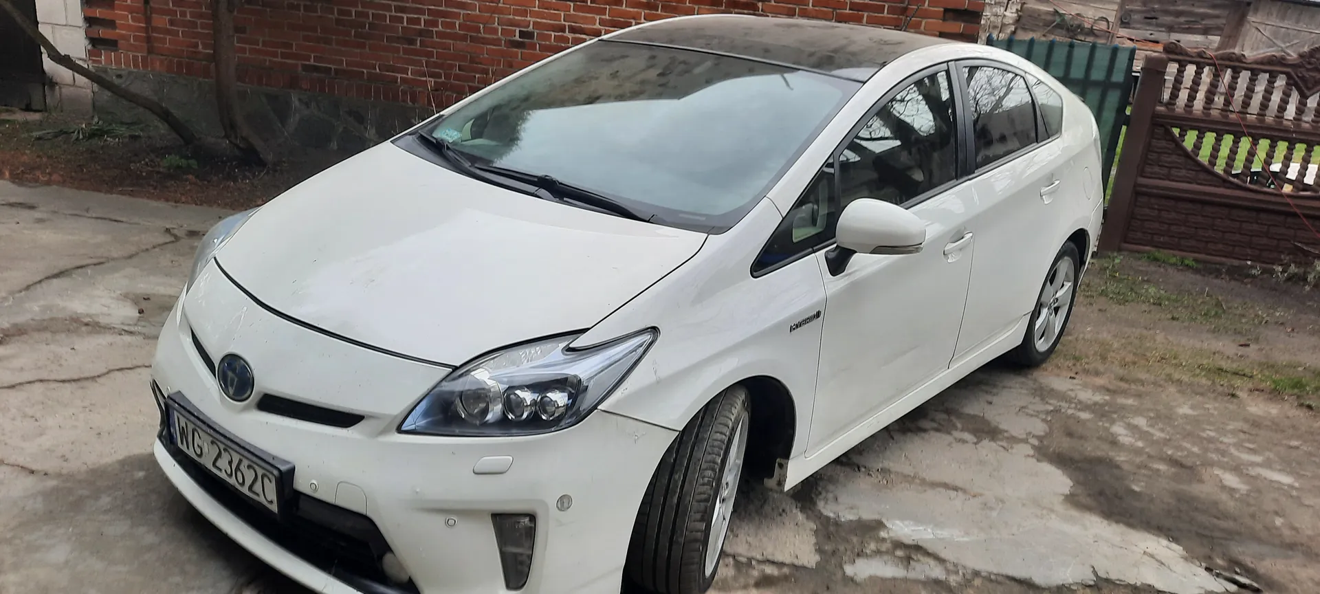 Biały samochód Toyota Prius z tablicą rejestracyjną WG-2362C zaparkowany na betonowej powierzchni. W tle ceglana ściana, zielony płot i brązowa drewniana brama. Na boku pojazdu widoczny napis 'Hybrid'.