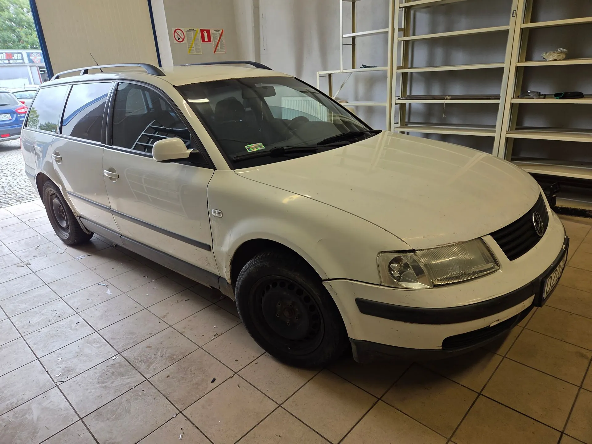 Biały samochód osobowy typu kombi zaparkowany w garażu lub warsztacie na płytkach ceramicznych. Pojazd stoi bokiem do widza, przodem w prawą stronę. W tle widoczne są białe regały magazynowe oraz znaki ostrzegawcze na ścianie.