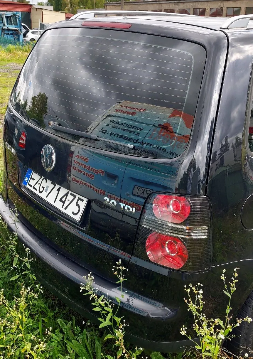 Tylny widok czarnego samochodu marki Volkswagen. Widoczna jest tylna szyba z ogrzewaniem, logo Volkswagena oraz tablica rejestracyjna o numerze EZG 71475. Na klapie bagażnika znajduje się oznaczenie 2.0 TDI. Samochód stoi na trawie, w tle widoczne są budynki przemysłowe.