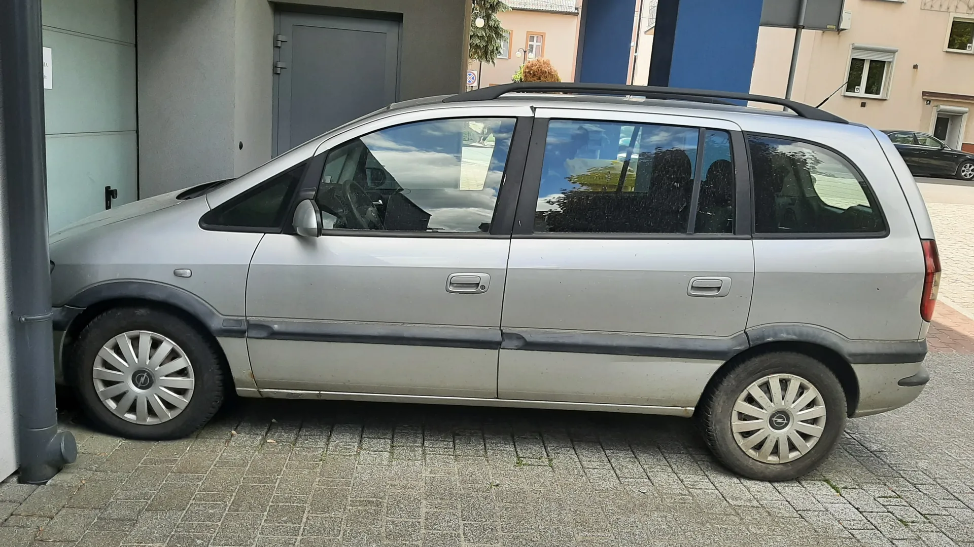 Srebrny samochód typu minivan zaparkowany na brukowanej powierzchni. Pojazd posiada czarny dachówkarz, przyciemniane szyby i srebrne felgi z logo w środku. W tle widoczne są budynki: szara brama, niebieska i beżowa elewacja z oknami oraz czarny samochód w oddali. Na lewo od minivana znajduje się szara rura i fragment szarej ściany z białą tabliczką.