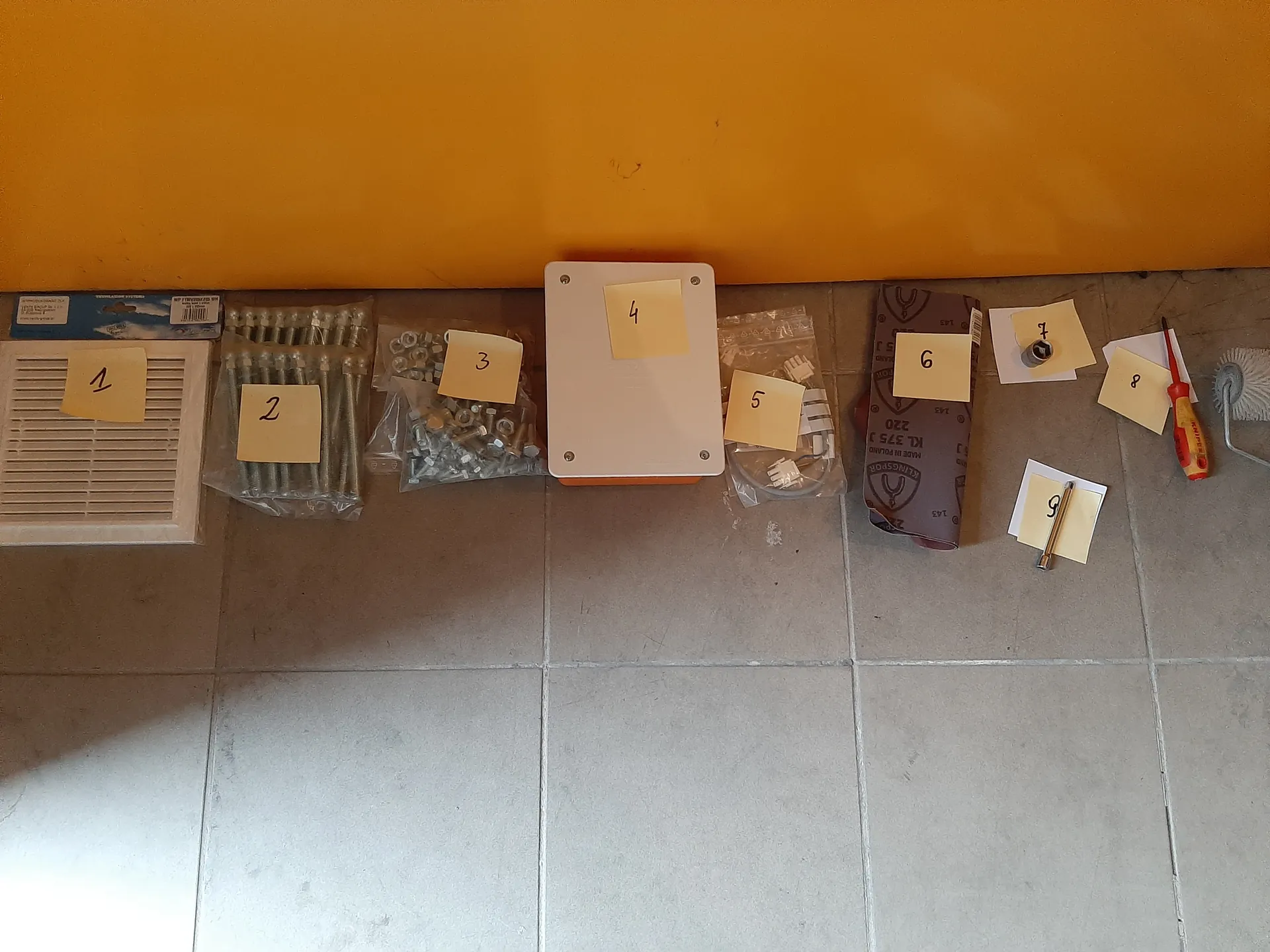 Na szarym płytkowym podłodze ułożono dziewięć przedmiotów oznaczonych żółtymi karteczkami z numerami od 1 do 9. Po lewej stronie znajduje się biała kratka wentylacyjna (1), obok opakowanie z metalowymi prętami (2), woreczek z nakrętkami i śrubami (3), białe prostokątne pudełko (4), woreczek z plastikowymi złączkami (5), arkusz papieru ściernego (6), mały metalowy element (7), czerwono-żółty śrubokręt (8) oraz metalowy przedmiot przypominający wiertło (9). W tle pomarańczowa ściana. Wszystkie numery na karteczkach są czytelne i ustawione pionowo.