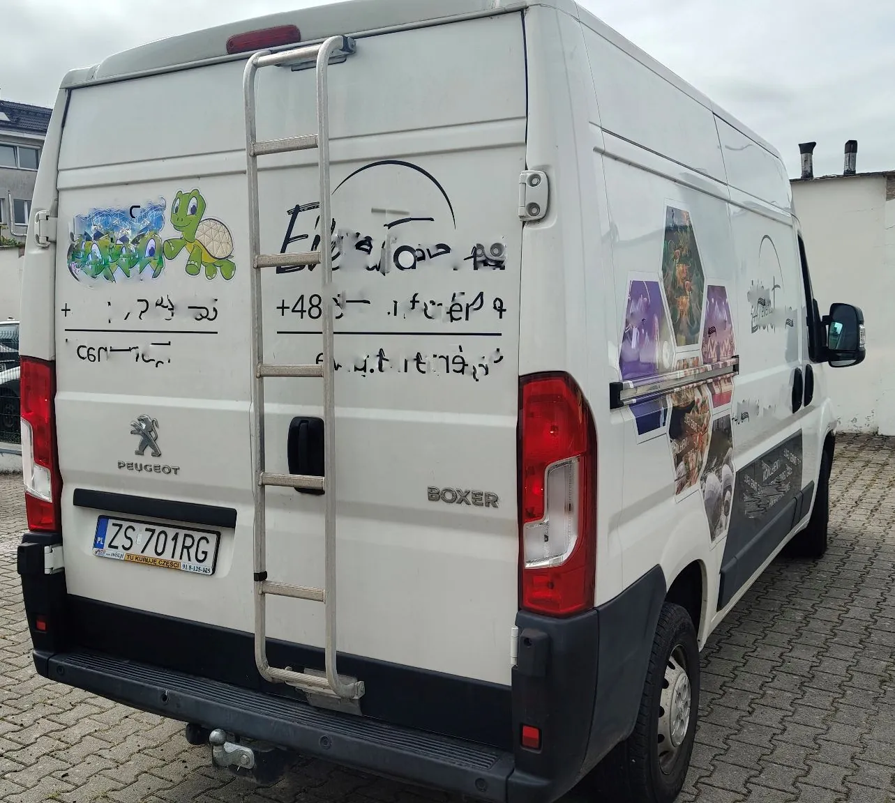 Biały samochód dostawczy marki Peugeot Boxer zaparkowany na kostce brukowej. Tył pojazdu posiada zamontowaną drabinkę, tablicę rejestracyjną o numerze ZS 701RG oraz napisy i rysunki naniesione na karoserię w formie graffiti. Na boku auta widoczne są kolorowe naklejki z grafikami.