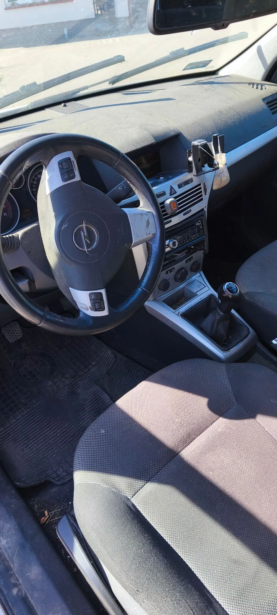 Wnętrze samochodu marki Opel, widok z perspektywy pasażera. Widoczna jest kierownica z logo producenta, deska rozdzielcza pokryta kurzem, konsola środkowa z radiem i lewarkiem zmiany biegów oraz ciemnoszare, materiałowe fotele. Przez przednią szybę widać fragment otoczenia w świetle dziennym.