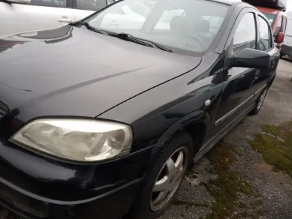 Czarny samochód osobowy typu sedan zaparkowany na podłożu z żwirem i trawą. Widoczna przód pojazdu z reflektorem, błyszcząca karoseria oraz fragmenty innych pojazdów na tle. Tło zawiera fragment białego i czerwonego pojazdu oraz ziemię z trawą.