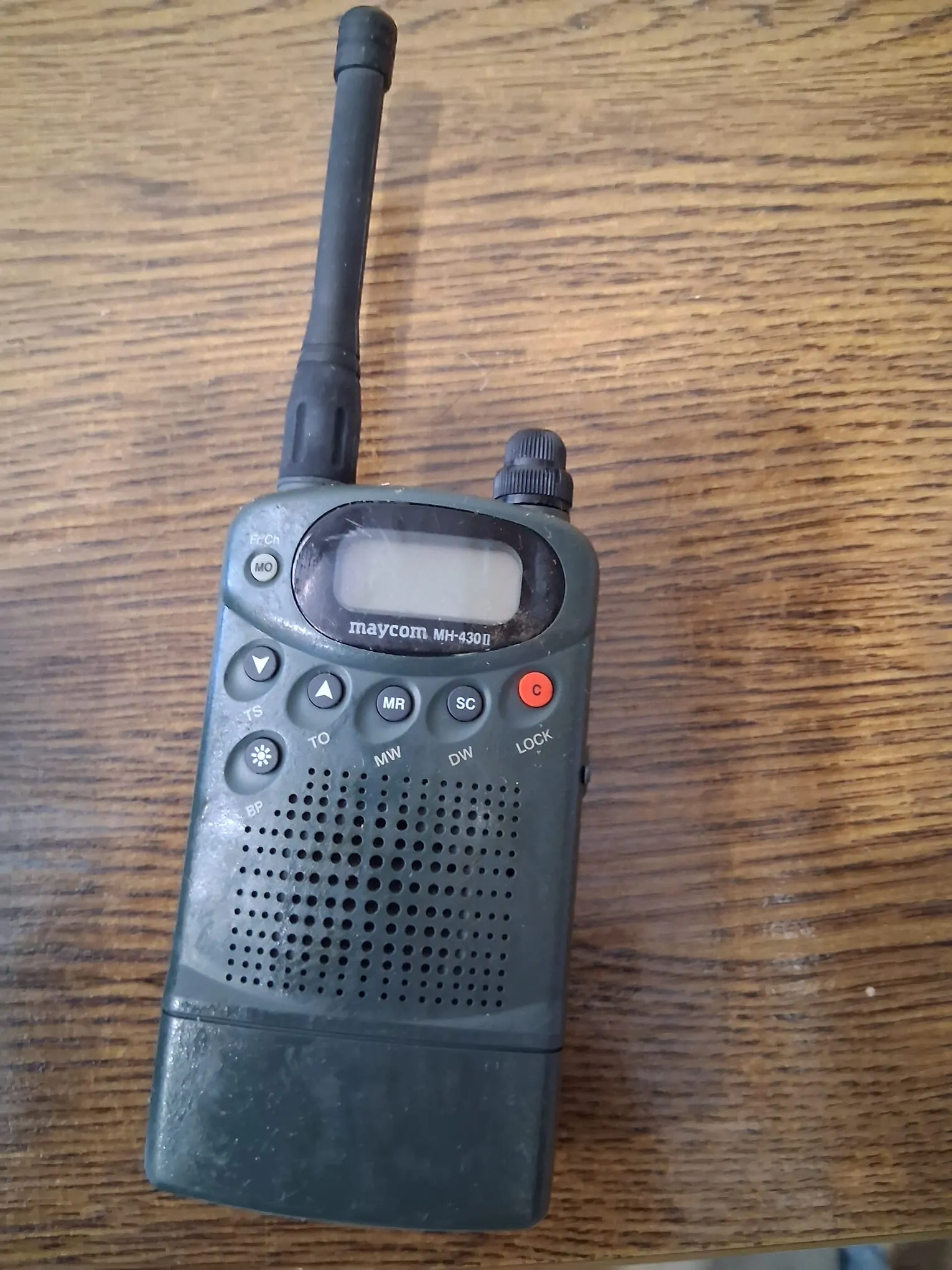 Ciemnoszara radiostacja przenośna marki Maycom, model MH-439II, leżąca na drewnianym blacie. Urządzenie posiada długą, czarną antenę na górze, mały ekran LCD oraz panel z przyciskami: czerwony przycisk w prawym górnym rogu oraz mniejsze przyciski z napisami MR, SC, TO, MW, DW i LOCK. W dolnej części znajduje się perforowany głośnik.