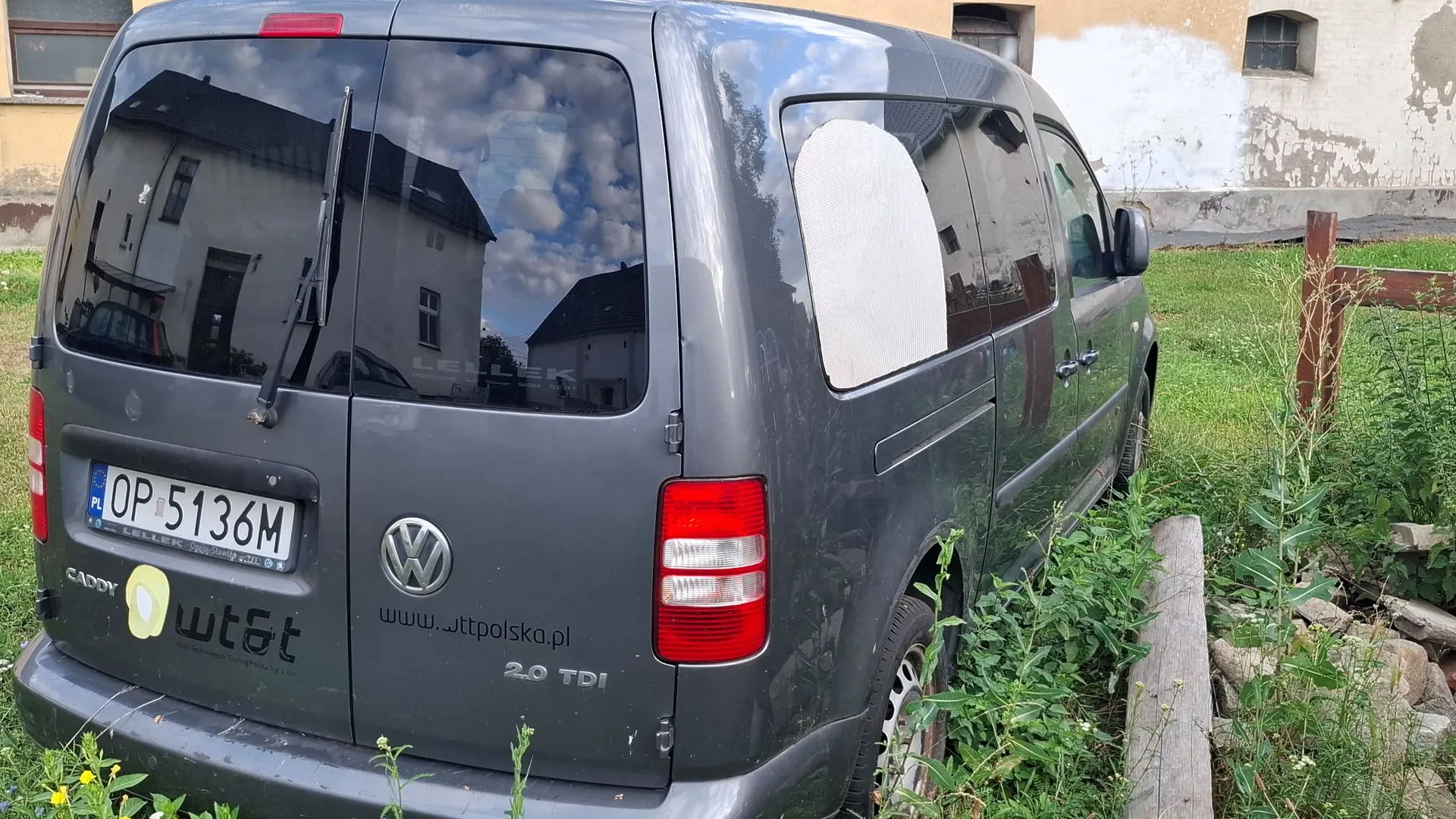 Tylna i boczna część szarego vana marki Volkswagen Caddy. Pojazd ma polską tablicę rejestracyjną o numerze OP 5136M. Na klapie bagażnika widoczne jest logo Volkswagena, napis 'CADDY', naklejka 'wt&t' oraz adres strony internetowej 'www.wttpolska.pl' i oznaczenie silnika '2.0 TDI'. Na bocznej szybie znajduje się biały, nieregularny kształt przypominający zaklejenie. Samochód zaparkowany jest na trawie obok betonowego krawężnika.