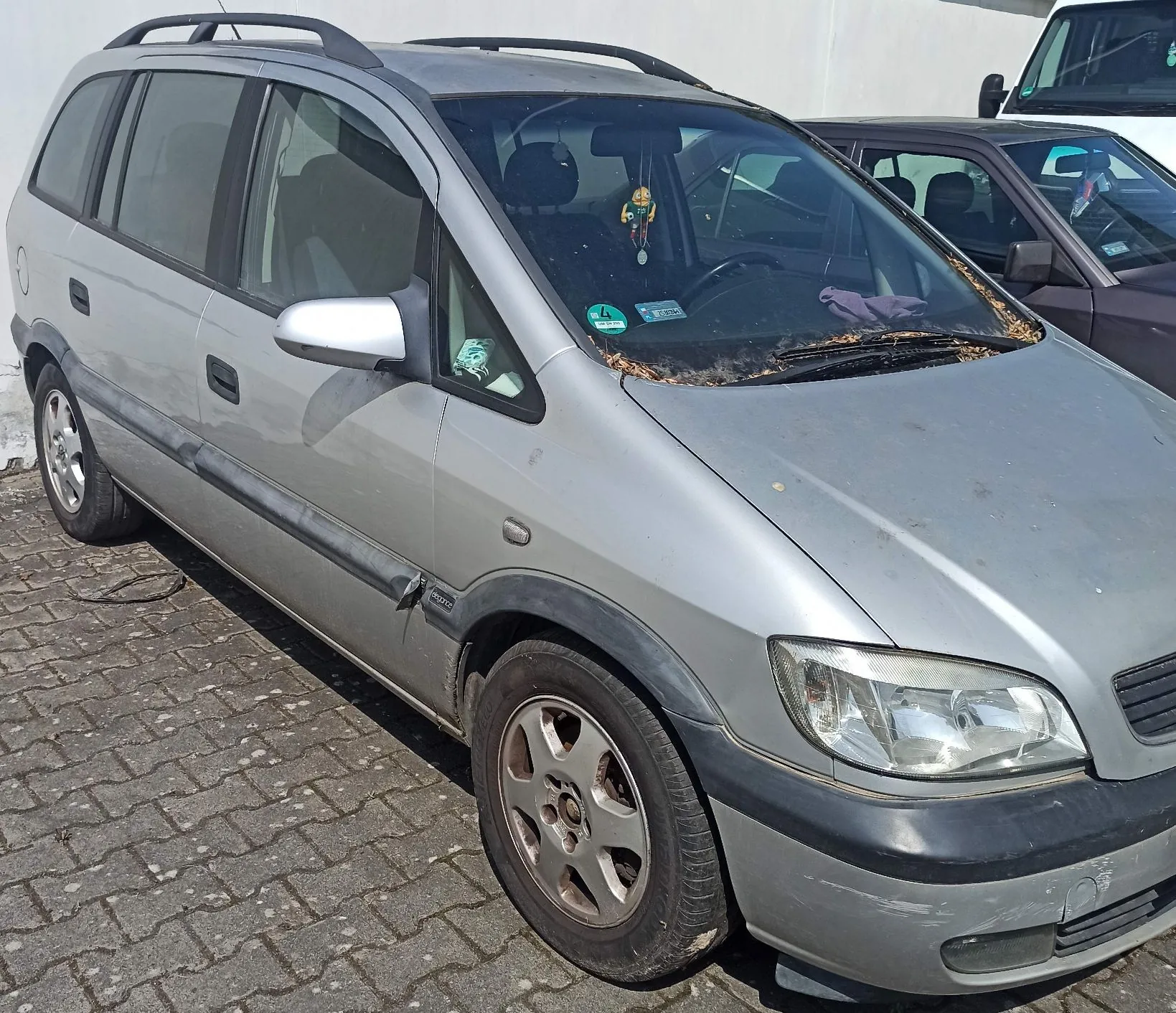 Srebrny samochód typu minivan zaparkowany na kostce brukowej. Pojazd ma czarne listwy boczne, relingi dachowe oraz przyciemniane tylne szyby. Na przedniej szybie widoczne są naklejki oraz zawieszona ozdoba. W tle widoczny jest fragment innego, ciemnego samochodu oraz biały pojazd dostawczy.