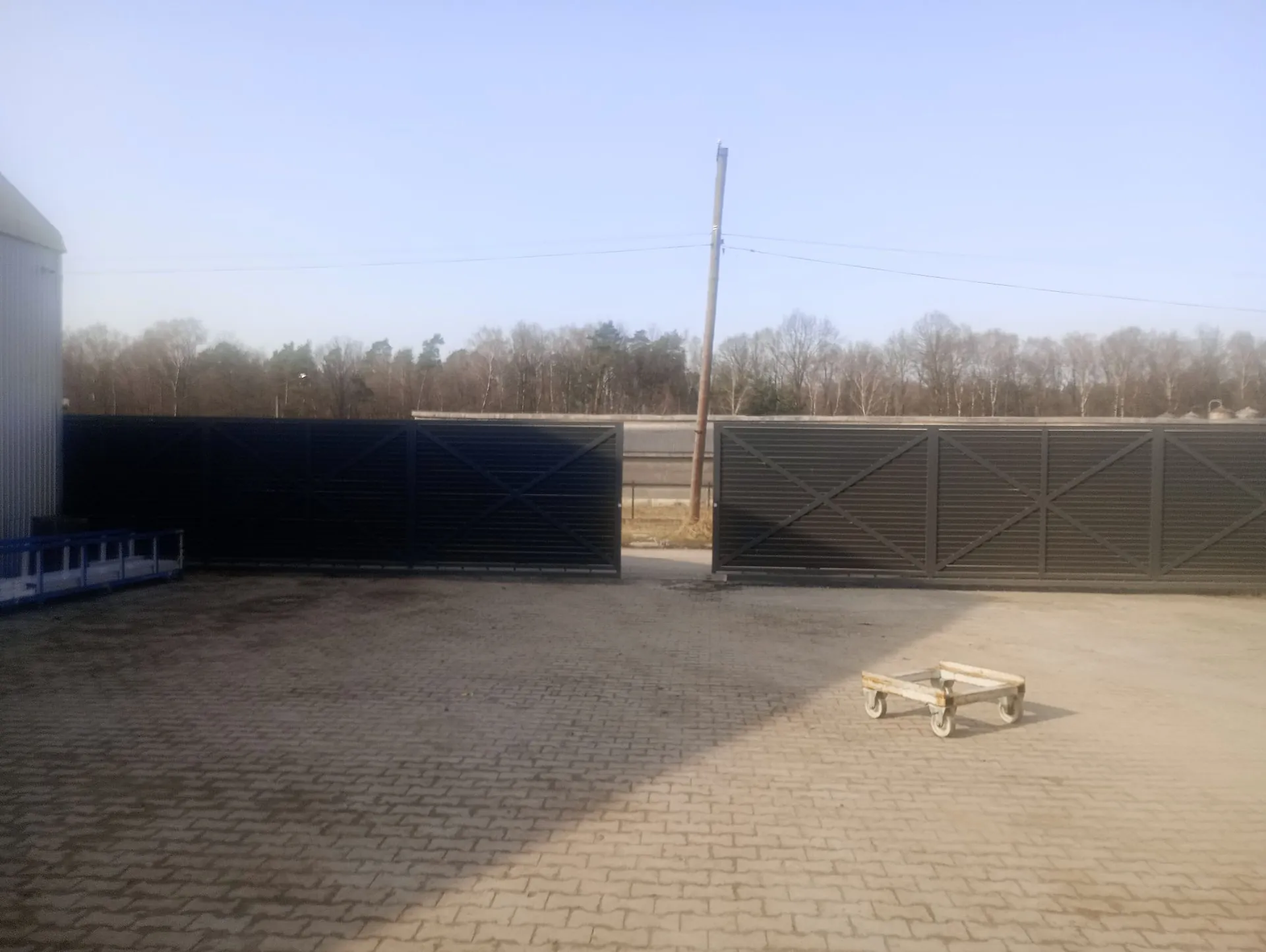 Zdjęcie przedstawia utwardzony plac z kostki brukowej, na którym znajduje się metalowy wózek transportowy na kółkach. W tle widoczny jest wysoki, ciemny płot z poziomych lameli, który jest częściowo otwarty. Za płotem znajduje się pas zieleni z bezlistnymi drzewami pod jasnym, bezchmurnym niebem. Po lewej stronie widoczny jest fragment białego budynku przemysłowego oraz niebieska barierka.