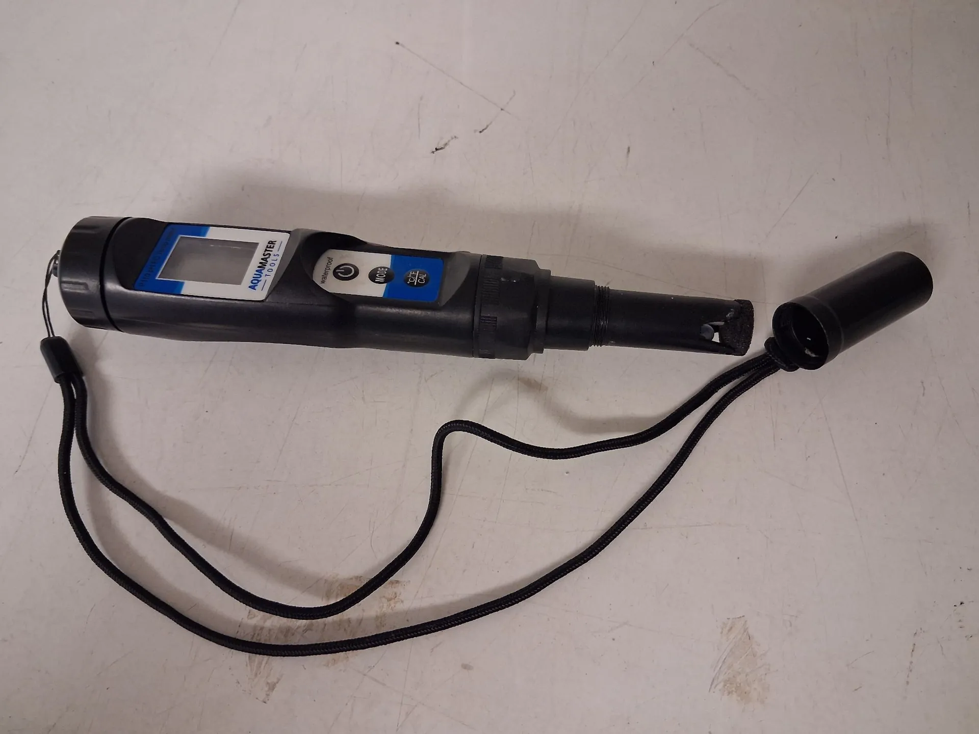 Czarny, przenośny miernik elektroniczny marki AquaMaster, leżący poziomo na białym tle. Urządzenie posiada niebieski wyświetlacz LCD oraz przyciski sterujące z napisami 'On/Off', 'Hold' oraz 'Cal'. Do obudowy przymocowany jest czarny sznurek. Po prawej stronie znajduje się odkręcona czarna zakrętka ochronna.