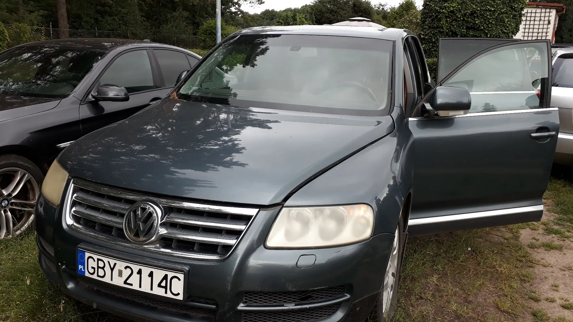Ciemnoszary samochód marki Volkswagen z otwartymi przednimi drzwiami po stronie pasażera. Pojazd posiada polską tablicę rejestracyjną o numerze GBY 2114C. W tle widoczny jest inny, czarny samochód oraz zieleń drzew i krzewów.