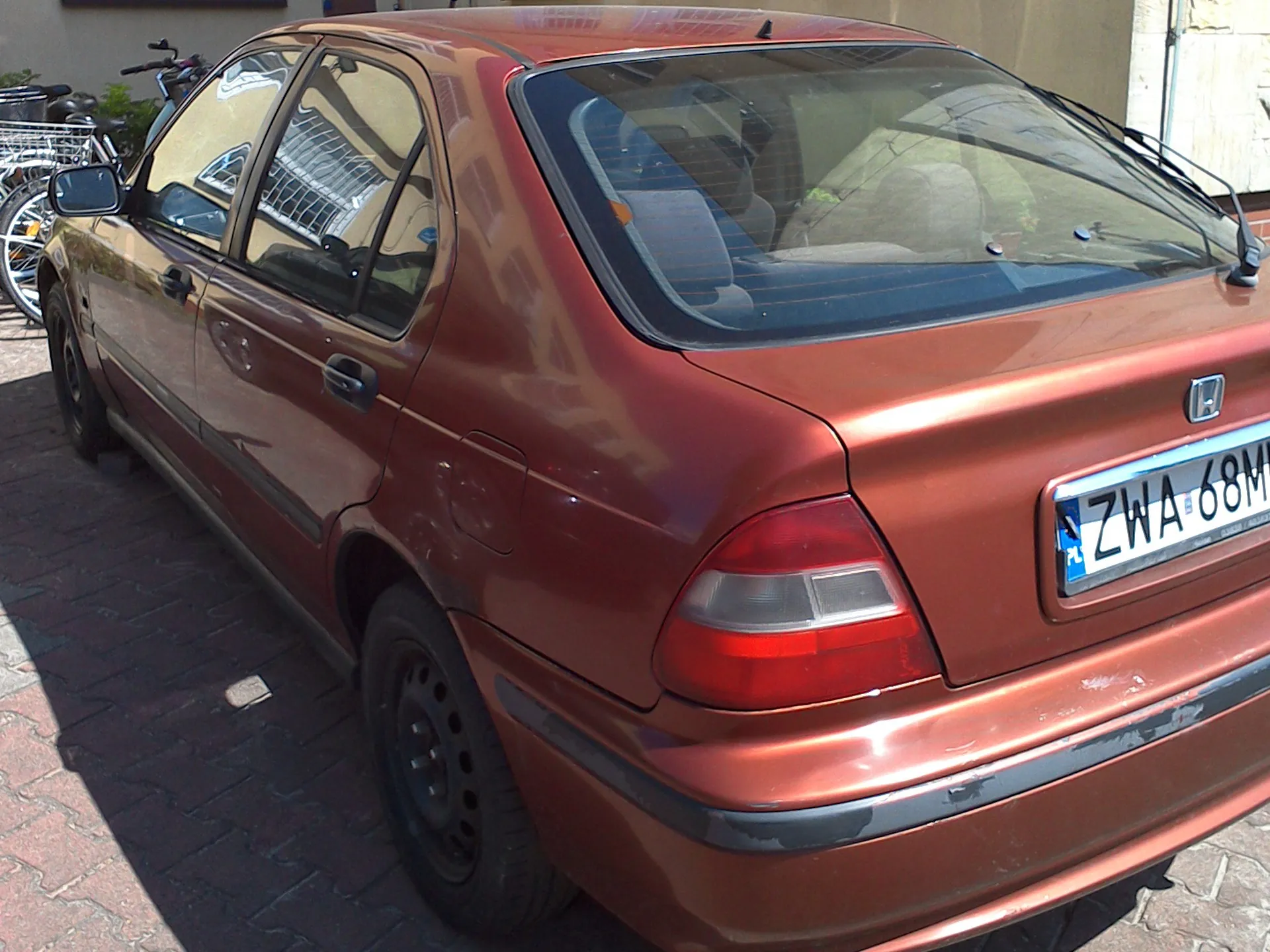 Widok z tyłu i z boku miedzianego samochodu osobowego marki Honda. Pojazd zaparkowany jest na kostce brukowej. Widoczna jest polska tablica rejestracyjna o numerze ZWA 68M.