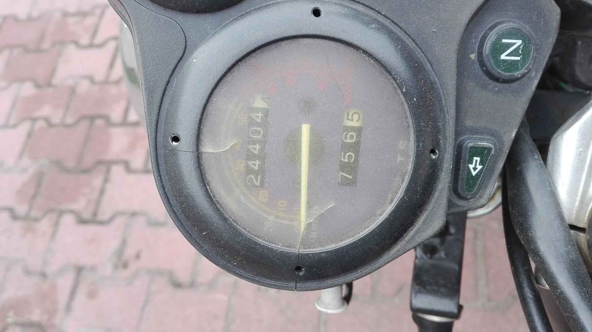 Zbliżenie na analogowy licznik przebiegu w motocyklu. Na tarczy widoczne są dwie liczby: 24404 oraz 7565. Obok licznika znajdują się dwa przyciski: górny z literą 'N' oraz dolny z ikoną strzałki skierowanej w dół. Całość osadzona jest w czarnej, plastikowej obudowie.