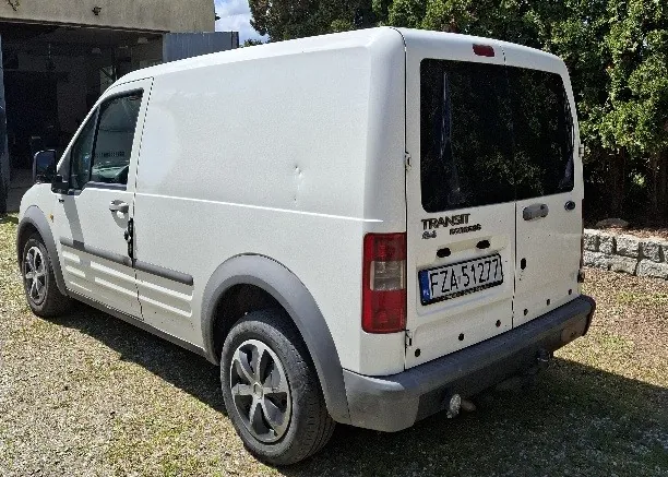 Biały samochód dostawczy Ford Transit z tablicą rejestracyjną FZA 5127 zaparkowany na żwirowym podwórku. W tle widoczne są drzewa, kamienna ściana oraz budynek z otwartą bramą garażową. Samochód ma czarne szyby tylne i srebrne felgi. Na tylnym zderzaku znajduje się napis TRANSIT.