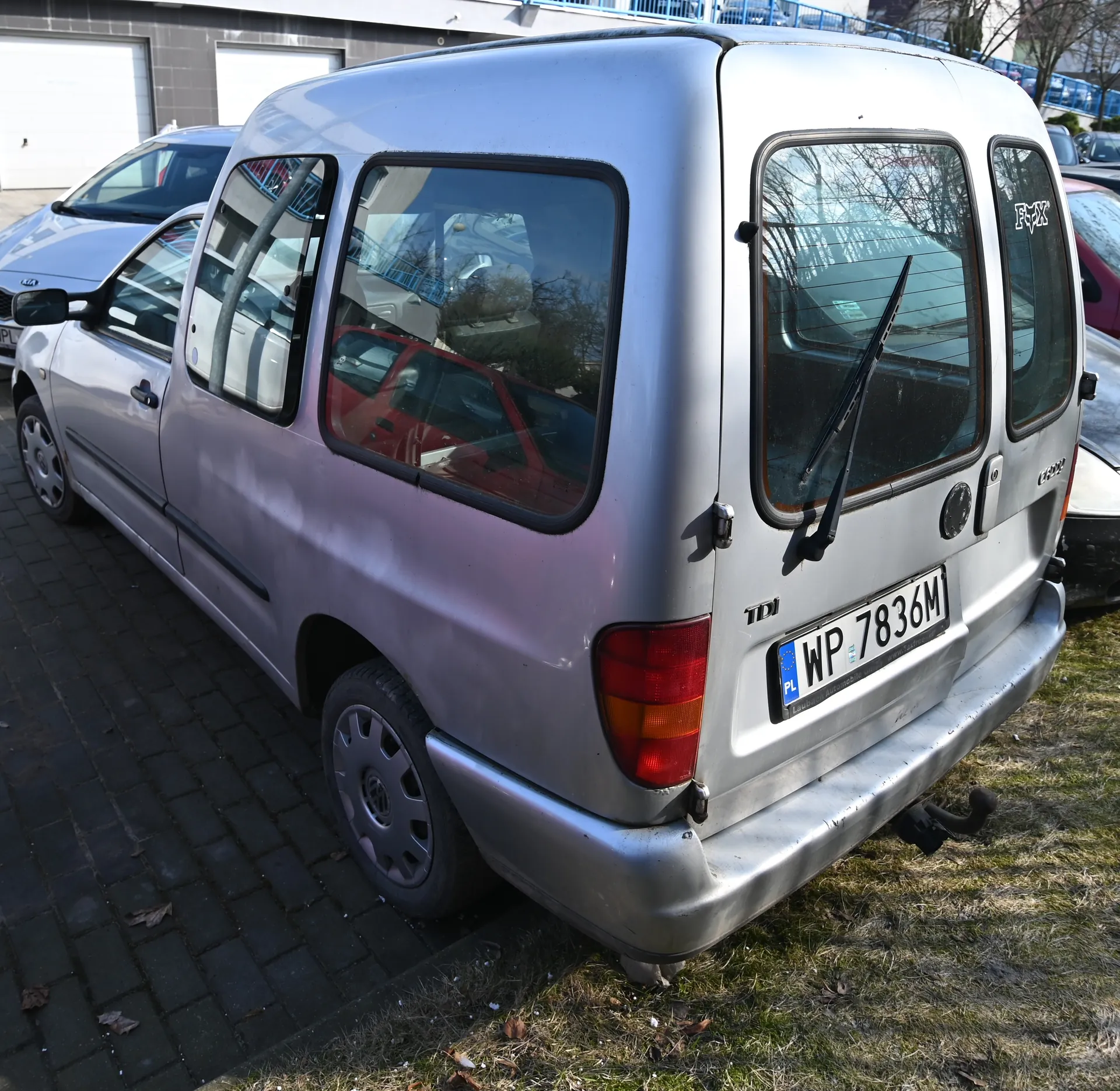 Srebrny samochód dostawczy z tyłu, z widoczną tablicą rejestracyjną WP 7836M oraz oznaczeniem TDI. Po lewej stronie widoczne są inne zaparkowane pojazdy, w tym srebrny samochód osobowy. Tło stanowią budynki z białymi bramami garażowymi i niebieskim ogrodzeniem. Samochód stoi na brukowanej powierzchni, obok trawnika z suchą trawą. Na tylnym szybie widoczny jest wycieraczka, a na bocznej szybie napis E-BOX.