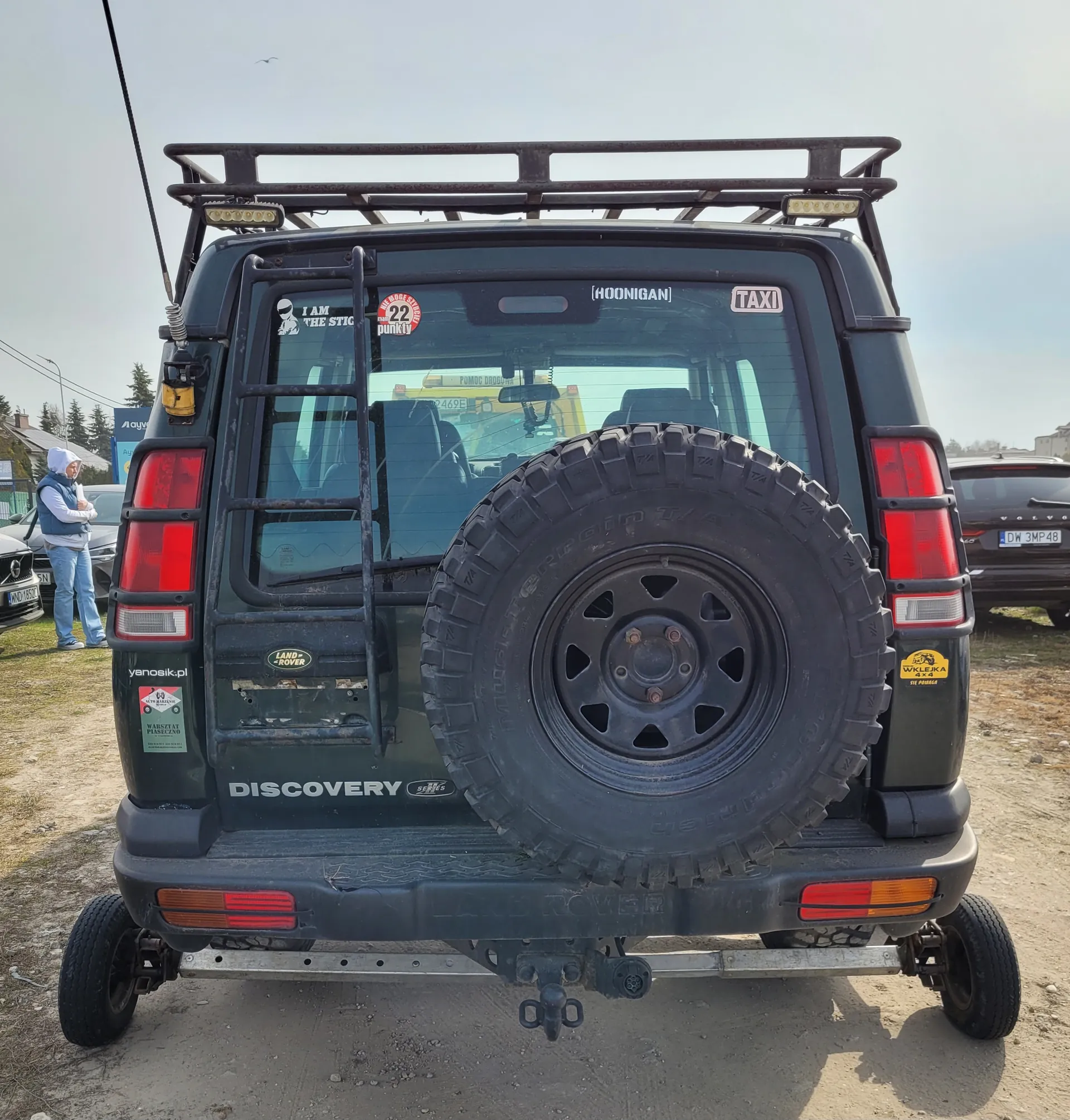 Tył ciemnego samochodu Land Rover Discovery z zamontowanym na drzwiach zapasowym kołem o grubym profilu. Na szybie tylnej naklejki: 'I AM THE STIG', czerwona z napisem '22 punkty', '[HOONIGAN]' oraz 'TAXI'. Na drzwiach widoczne napisy 'DISCOVERY', 'LAND ROVER' i 'yanosik.pl', a po prawej stronie żółta naklejka 'WYKŁADKA'. Po lewej stronie samochodu stoi osoba w białej bluzie i niebieskiej kamizelce. W tle widać inne pojazdy, w tym samochód z tablicą rejestracyjną 'DW 3MP48', oraz budynki i drzewa na tle jasnego nieba. Samochód wyposażony w dachowy bagażnik i drabinkę po lewej stronie.