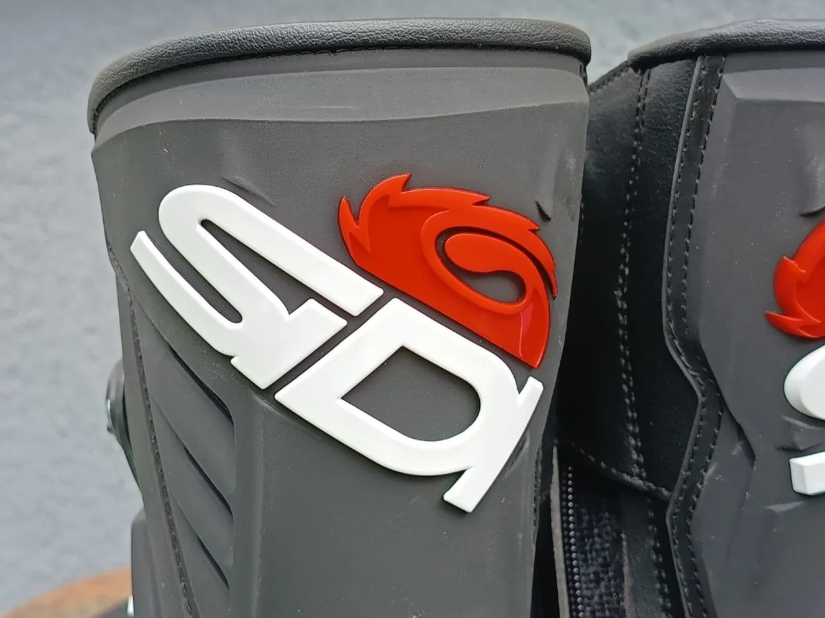 Zbliżenie na ciemnoszary element ochronny buta motocyklowego. Na powierzchni znajduje się biały, wypukły napis 'SD' oraz czerwony, wypukły symbol przypominający stylizowany płomień lub głowę ptaka.