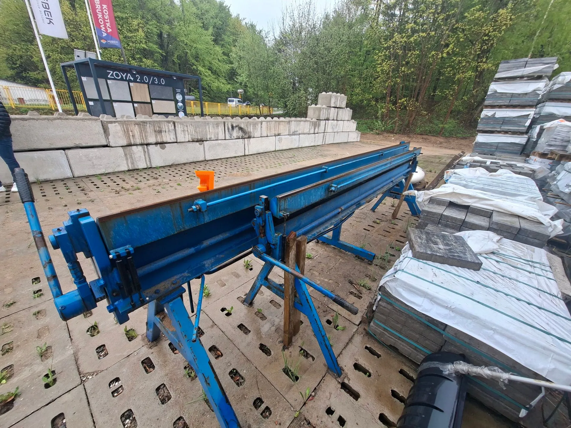 Niebieska, przemysłowa przecinarka do kostki brukowej ustawiona na betonowym placu. Maszyna posiada długą prowadnicę i mechanizm korbowy po lewej stronie. W tle widoczne są stosy betonowych krawężników, palety z kostką brukową przykryte białą folią oraz tablica informacyjna z napisem 'ZOYA 2.0/3.0'. W oddali znajduje się pas zieleni z drzewami.