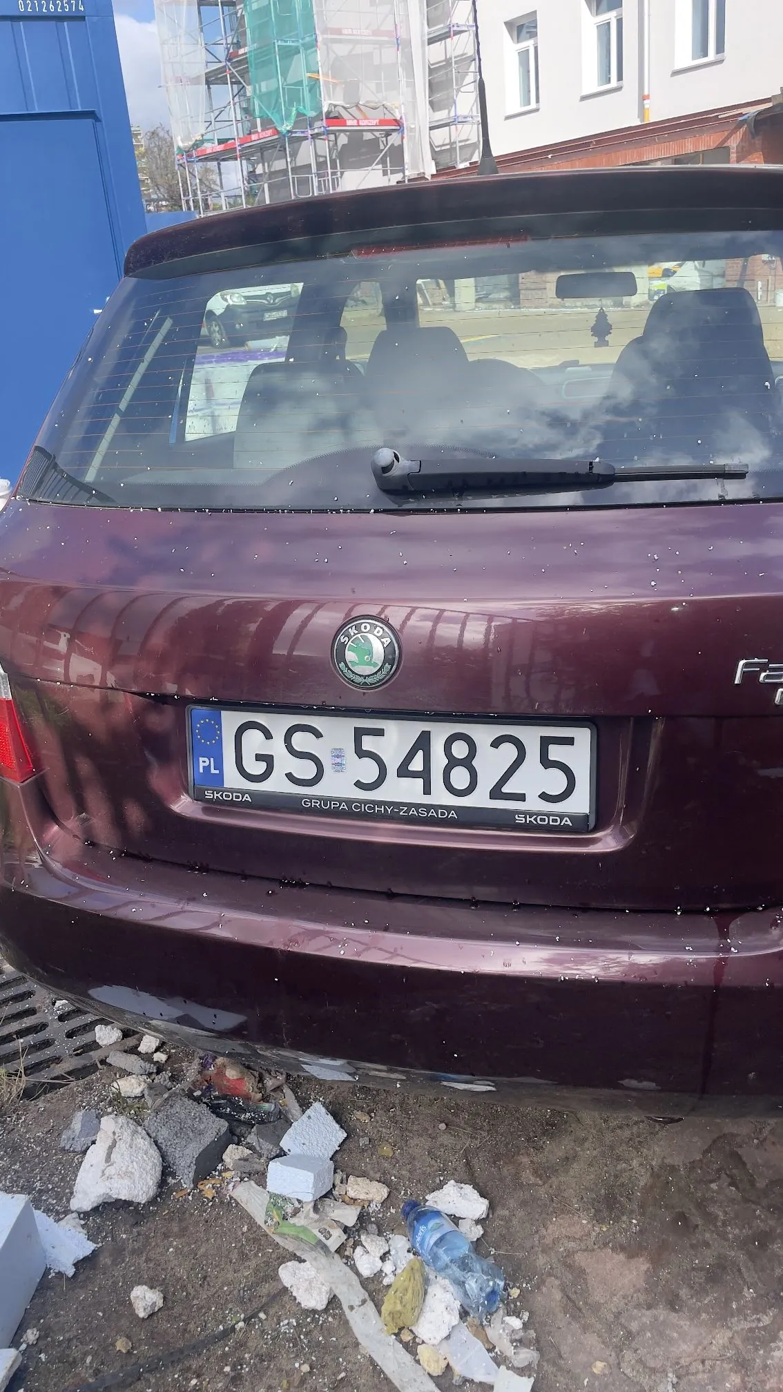 Tylny widok bordowego samochodu marki Skoda z polską tablicą rejestracyjną o numerze GS 54825. Pod samochodem widoczne są gruzy, białe kawałki styropianu oraz plastikowa butelka z wodą. W tle widać budynek w trakcie remontu z rusztowaniami.