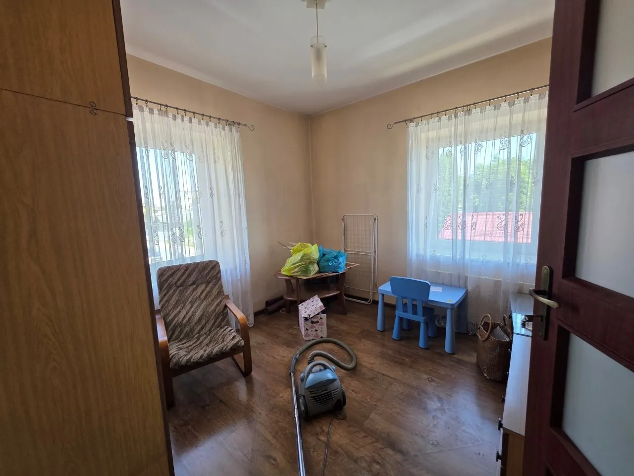 Wnętrze pokoju z drewnianą podłogą i beżowymi ścianami. W pomieszczeniu znajdują się dwa okna z białymi, firankami, brązowy fotel z wzorzystym obiciem, mały niebieski stolik z krzesełkiem dla dziecka oraz drewniany stolik z kolorowymi torbami na górze. Na podłodze leży szary odkurzacz z rurą, a obok niego wiklinowy kosz. Po lewej stronie widoczny jest fragment drewnianej szafy, a po prawej otwarta brązowa drzwi.