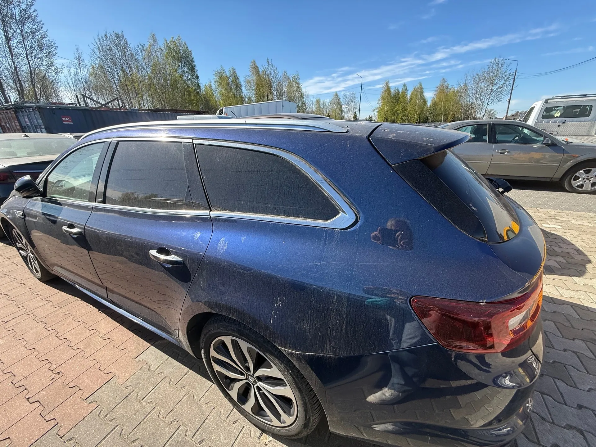 Niebieski samochód typu kombi z dachówką, zaparkowany na brukowanym placu. Widoczne są brud na karoserii, felgi aluminiowe oraz refleksja osoby fotografującej na tylnej szybie. W tle: srebrny sedan, biały van, drzewa, niebo oraz kontener z napisem "TAL". Inne pojazdy i elementy otoczenia są ustawione zgodnie z horyzontem.