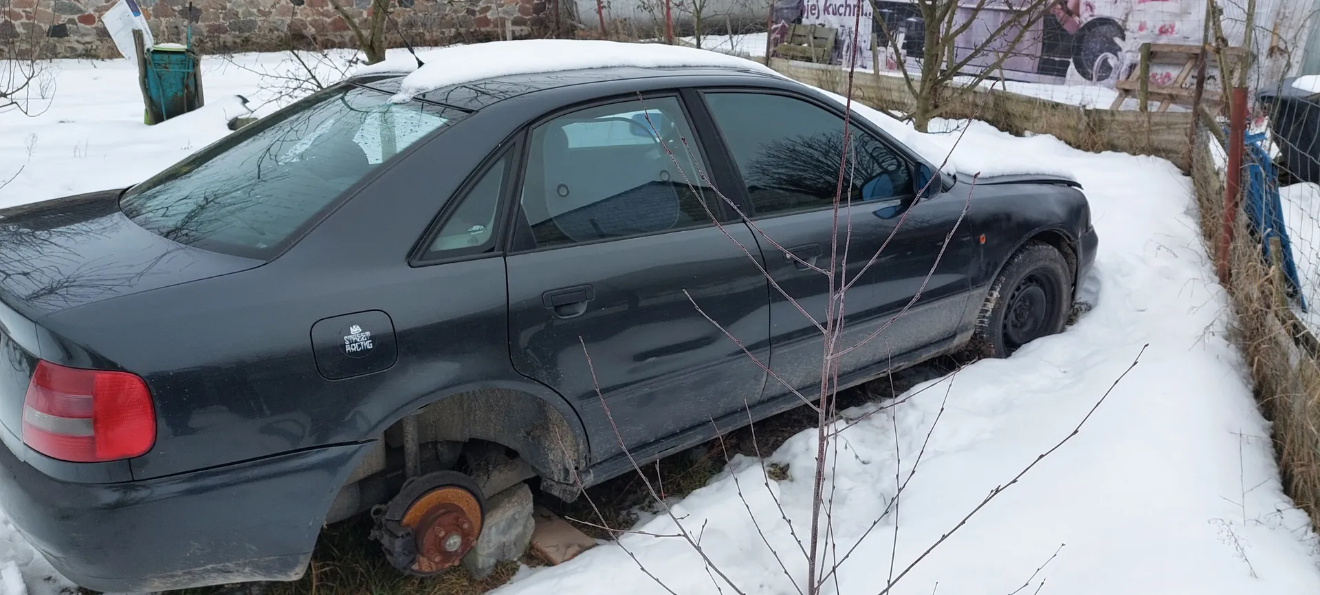 Czarny samochód osobowy typu sedan zaparkowany na śniegu. Na dachu pojazdu leży warstwa śniegu. Brakuje lewego tylnego koła, a jego miejsce zajmuje kamienny blok. Na klapie bagażnika widoczny jest naklejony napis 'STREET RACING' z logo. W tle: kamienna ściana, niebieski pojemnik, nagie drzewa, ogrodzenie z siatki oraz fragment tablicy z niewyraźnym napisem 'kuchni...'. Śnieg pokrywa teren wokół samochodu.