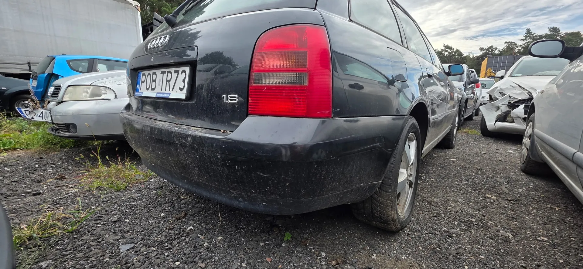 Tylna część czarnego samochodu marki Audi z tablicą rejestracyjną POB TR73 oraz oznaczeniem modelu 1.8. Samochód znajduje się na placu z żwirem obok innych uszkodzonych pojazdów, w tym srebrnego samochodu po lewej i białego z rozbitym przodem po prawej.