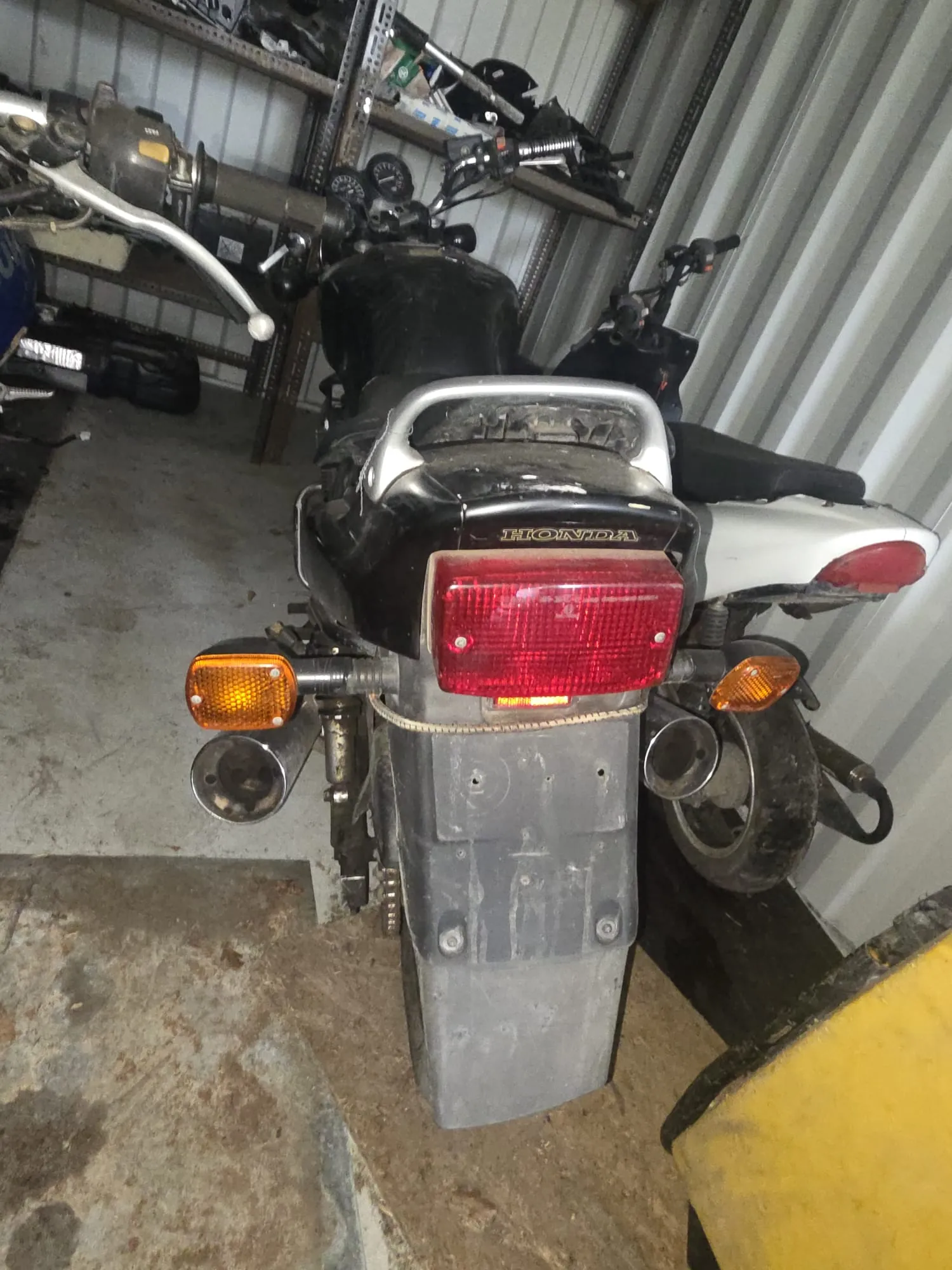 Z tyłu widok motocykla marki Honda w garażu. Czerwone światło tylne, pomarańczowe kierunkowskazy po obu stronach, szara tablica rejestracyjna bez widocznego numeru. Na bagażniku widoczny napis HONDA. W tle metalowe półki z narzędziami i częściami, betonowa podłoga z plamami. Po prawej stronie fragment żółtego obiektu.