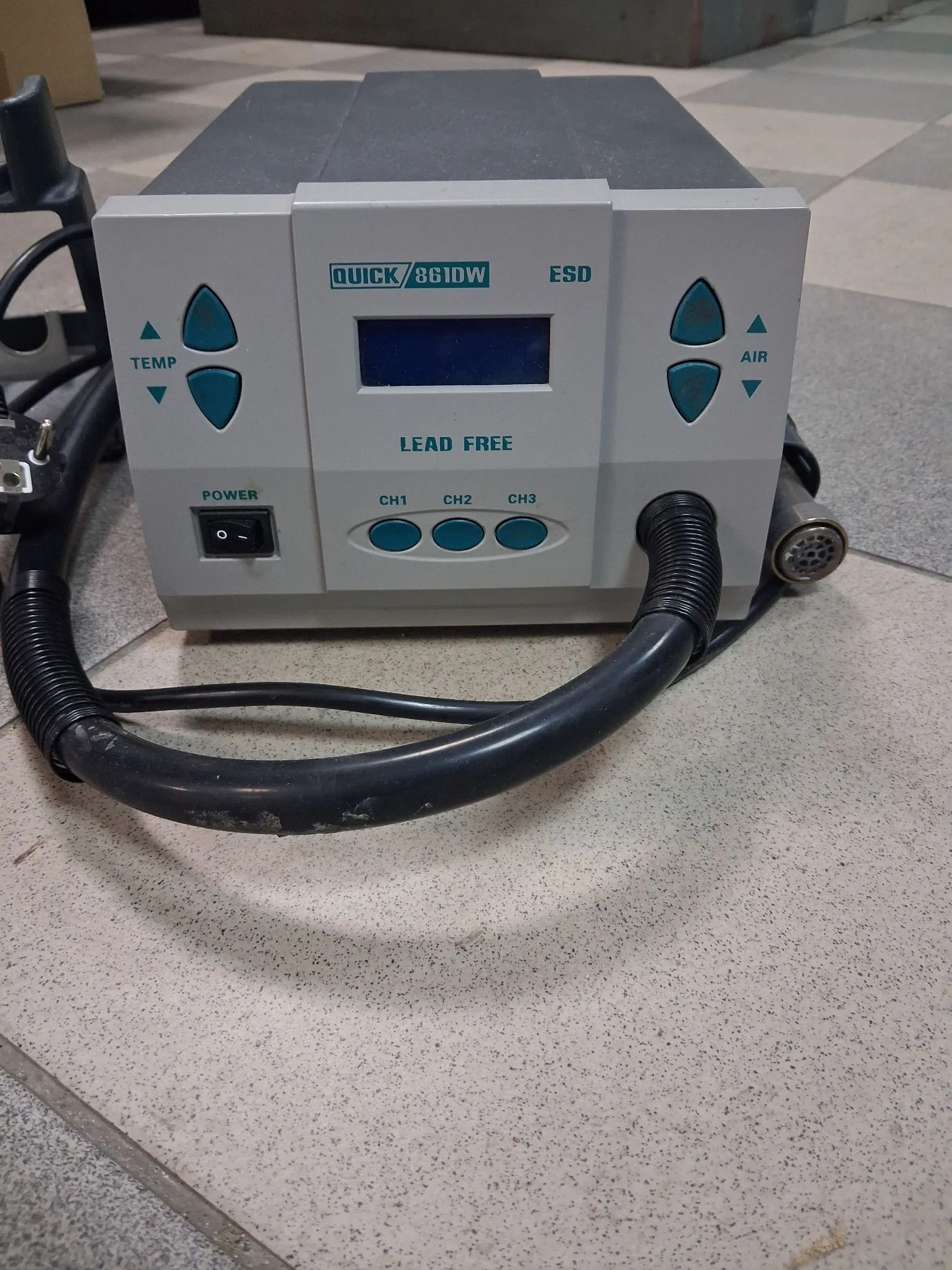Urządzenie elektroniczne, prawdopodobnie stacja lutownicza, w kolorze jasnoszarym. Na przednim panelu znajduje się niebieski wyświetlacz cyfrowy, napisy 'QUICK 861DW', 'ESD' oraz 'LEAD FREE'. Po lewej stronie są przyciski regulacji temperatury ('TEMP'), po prawej regulacji przepływu powietrza ('AIR'). Na dole znajdują się trzy przyciski wyboru kanałów (CH1, CH2, CH3) oraz przełącznik zasilania ('POWER'). Do urządzenia podłączone są czarne kable i przewody w peszlach.