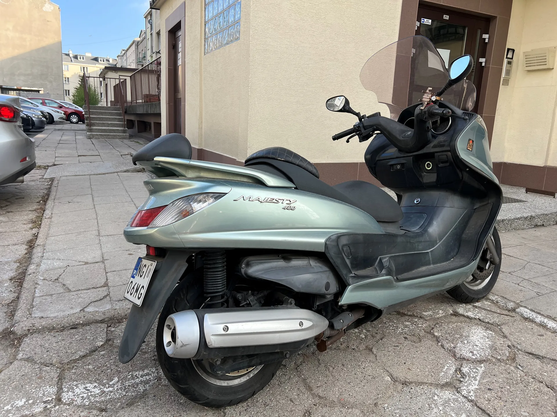 Srebrny skuter Honda Majesty 400 zaparkowany na brukowanej ulicy. Widoczna tablica rejestracyjna z numerem NGI 06MG. W tle budynki mieszkalne, schody, metalowe poręcze oraz zaparkowane samochody. Skuter ma czarne siedzenie, lusterka boczne i przezroczystą osłonę przednią. Tekst MAJESTY 400 umieszczony na boku skutera.