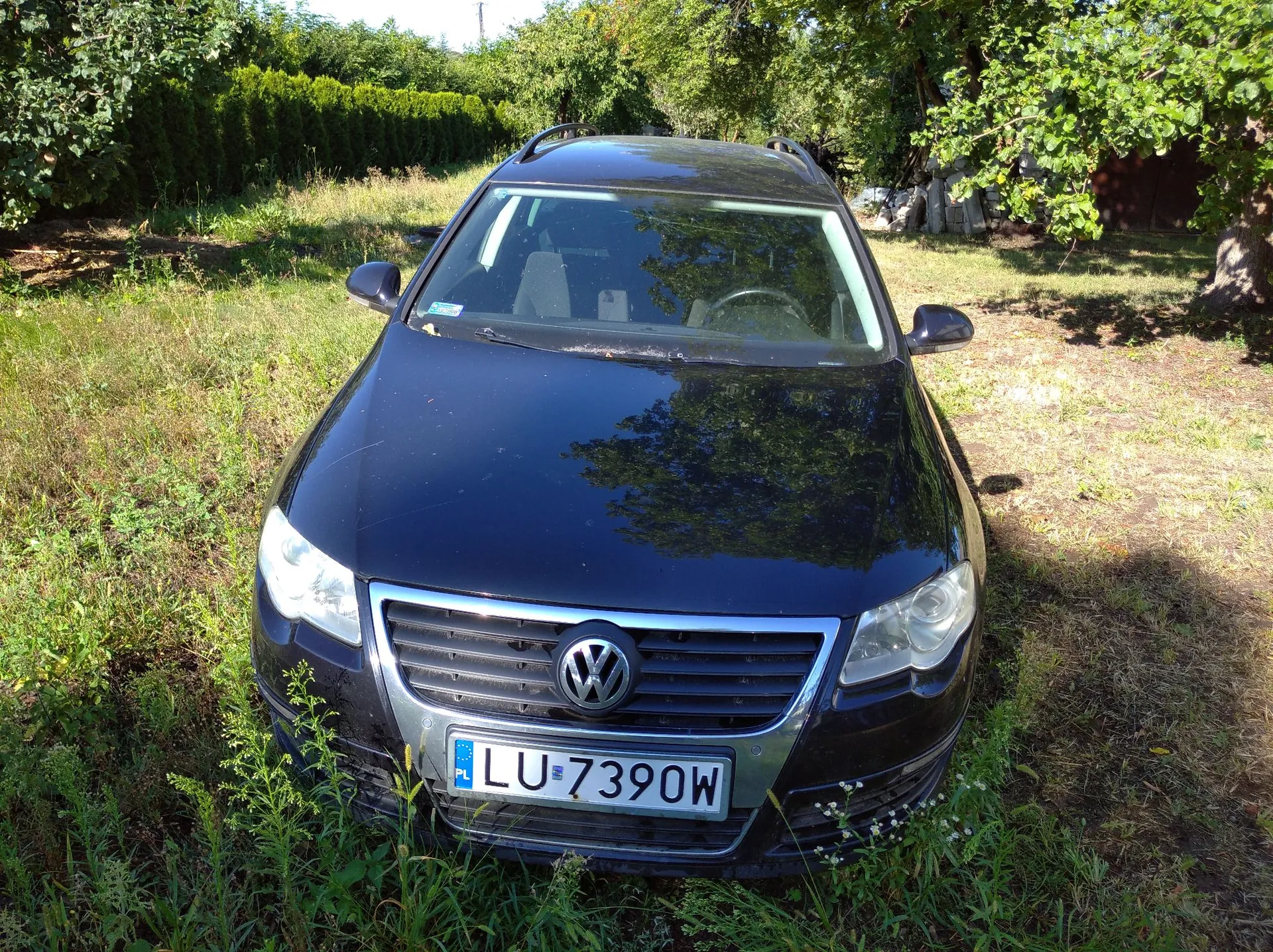 Czarny samochód marki Volkswagen zaparkowany na trawiastym terenie w otoczeniu zieleni i drzew. Widoczny jest przód pojazdu z tablicą rejestracyjną o numerze LU 7390W.