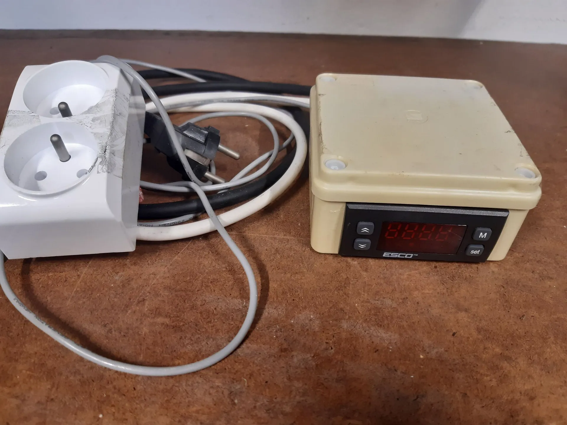Zestaw urządzeń elektrycznych leżących na brązowej, drewnianej powierzchni. Po lewej stronie znajduje się biała rozgałęźnik elektryczny z dwoma gniazdami, do którego podłączone są kable. Po prawej stronie znajduje się beżowa plastikowa obudowa z cyfrowym wyświetlaczem LED i przyciskami sterującymi. Na obudowie widnieje napis 'ESCO' oraz oznaczenia przycisków 'M' i 'set'.