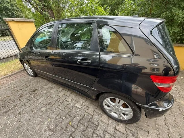 Czarny samochód osobowy typu hatchback zaparkowany na kostce brukowej. Pojazd jest pokazany z profilu od strony pasażera. W tle widoczny jest żółty mur, czarny metalowy płot oraz zielone drzewa i krzewy.