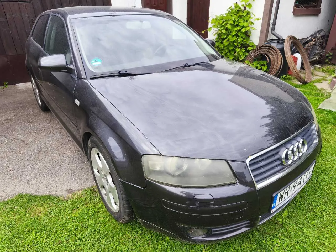 Czarny samochód marki Audi zaparkowany na trawie przed budynkiem. Widoczny jest przód i lewy bok pojazdu. Samochód ma polską tablicę rejestracyjną o numerze WR 941GE.