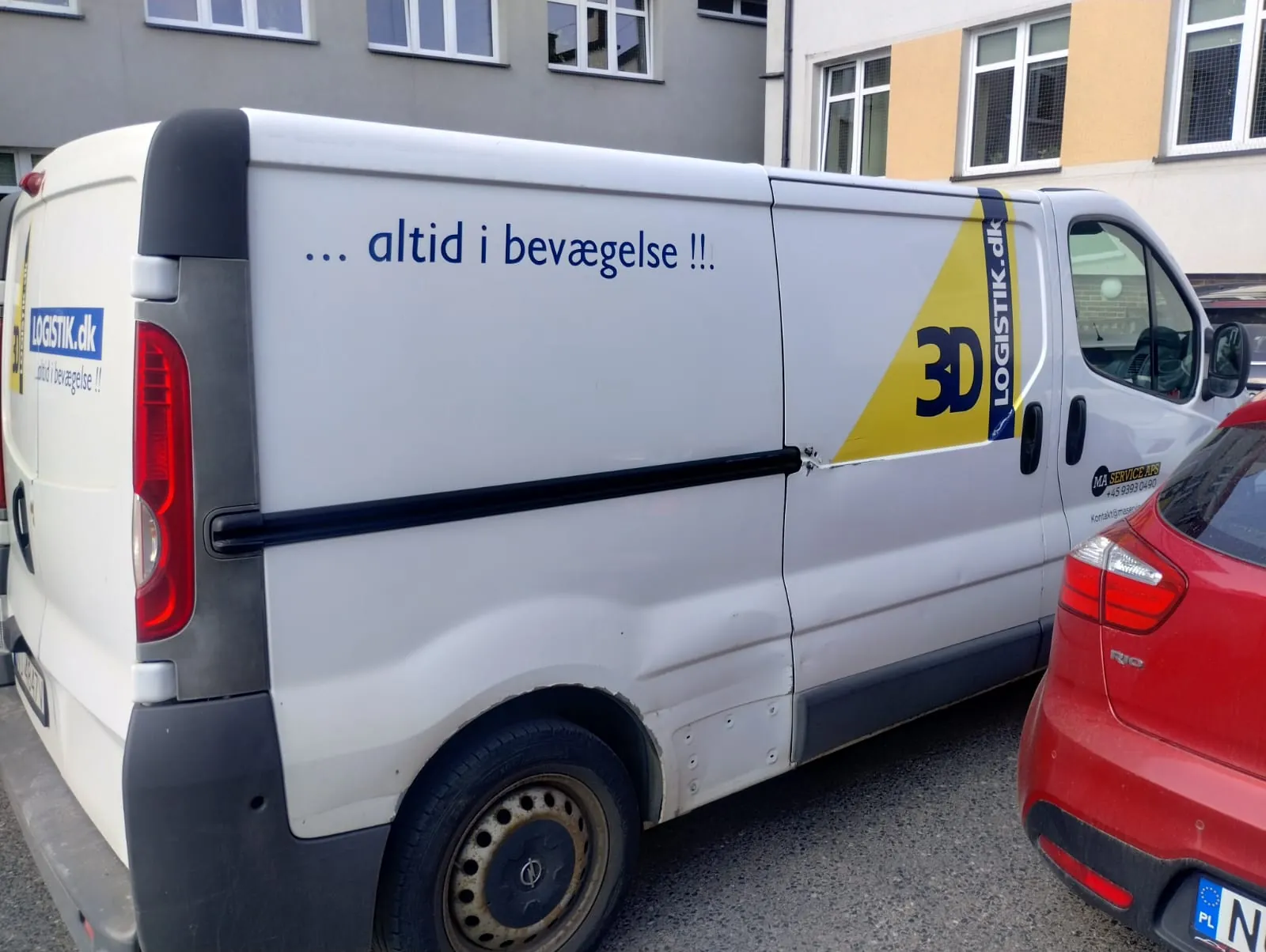 Biały furgon z napisem "... altid i bevægelse !!" na boku, logo "LOGISTIK.dk" na tylnej i bocznej części, żółty trójkąt z napisem "3D" oraz pionowym logo "LOGISTIK.dk". Widoczne są również czerwone tylne światła, czarne elementy nadwozia i koło z brudnym felgiem. Obok furgonu stoi czerwony samochód z częściowo widoczną tablicą rejestracyjną. W tle budynki z oknami.