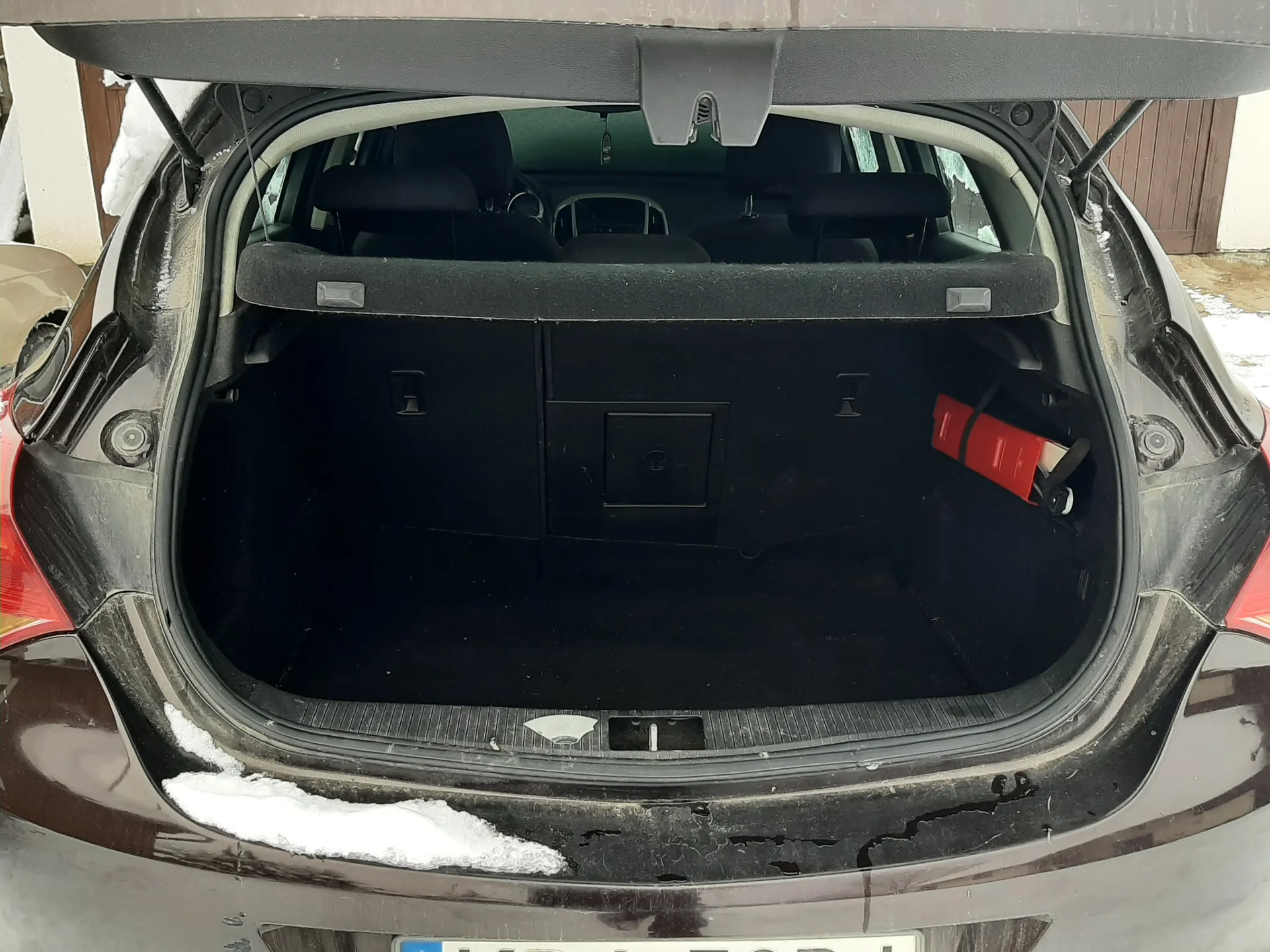Otwarty bagażnik ciemnego samochodu z widocznym czerwonym gaśnikiem po prawej stronie. Na krawędziach bagażnika śnieg, w tle budynek z brązowymi drzwiami i śniegiem na ziemi. Widoczne siedzenia tylne i częściowo tablica rejestracyjna.
