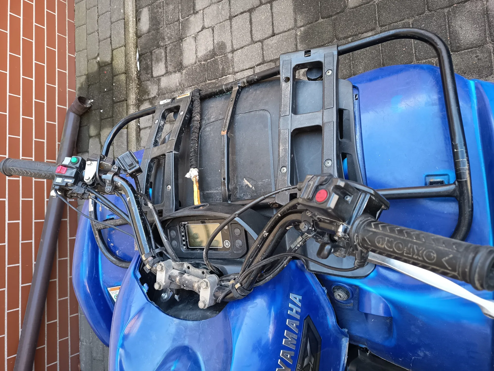 Niebieski motocykl marki YAMAHA z widocznym wyświetlaczem cyfrowym, czarnymi uchwytami kierownicy z napisem OXFORD oraz czarnym bagażnikiem. W tle ceglana ściana i brukowana powierzchnia. Na boku zbiornika widoczny napis YAMAHA.