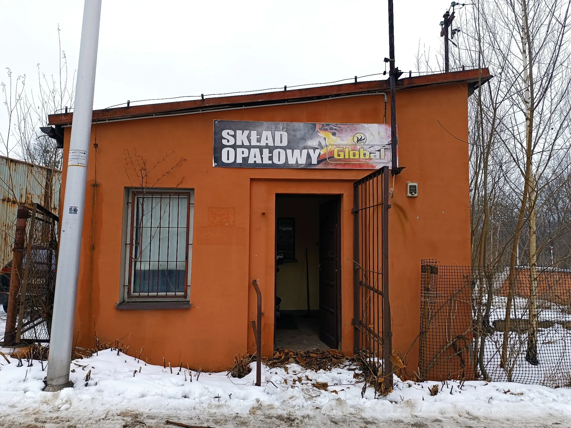 Pomarańczowy, parterowy budynek z napisem na szyldzie: 'SKŁAD OPAŁOWY Global'. Budynek posiada jedno okno z kratami oraz otwarte wejście prowadzące do ciemnego wnętrza. Przed budynkiem leży śnieg, a po prawej stronie znajduje się metalowy płot i bezlistne drzewa. Po lewej stronie widoczny jest biały słup.